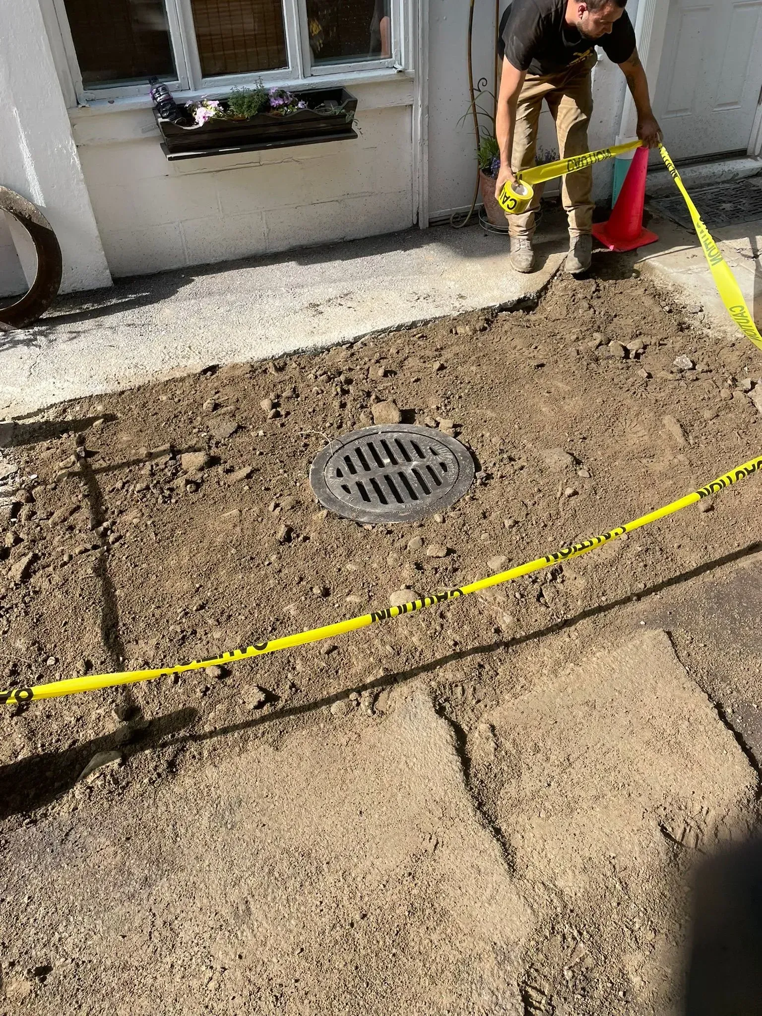 Worker using a yellow tape measure around a circular drain on a concrete patio near a building.