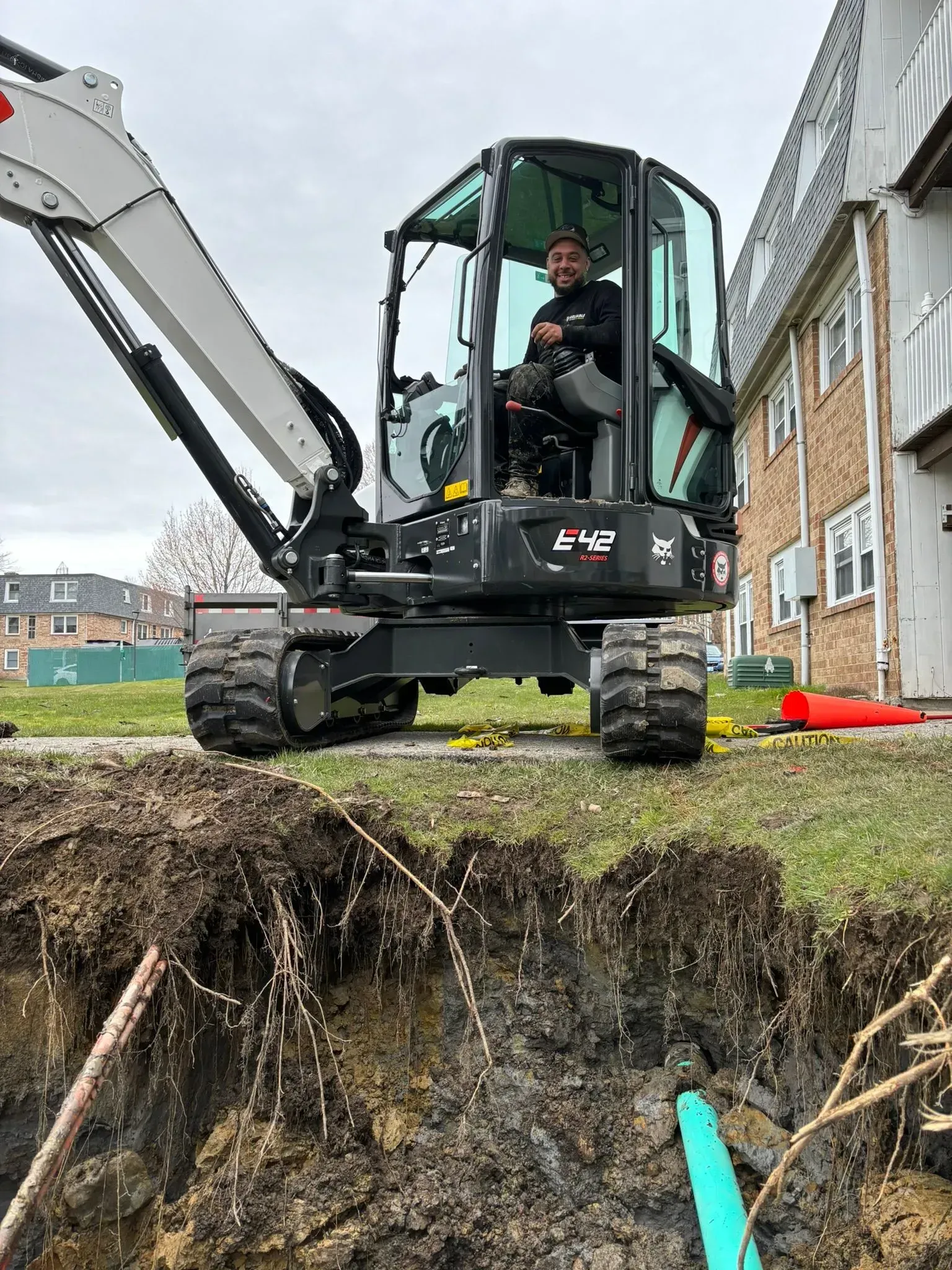 Excavator on a grassy excavation edge beside houses, with exposed roots and pipes below.