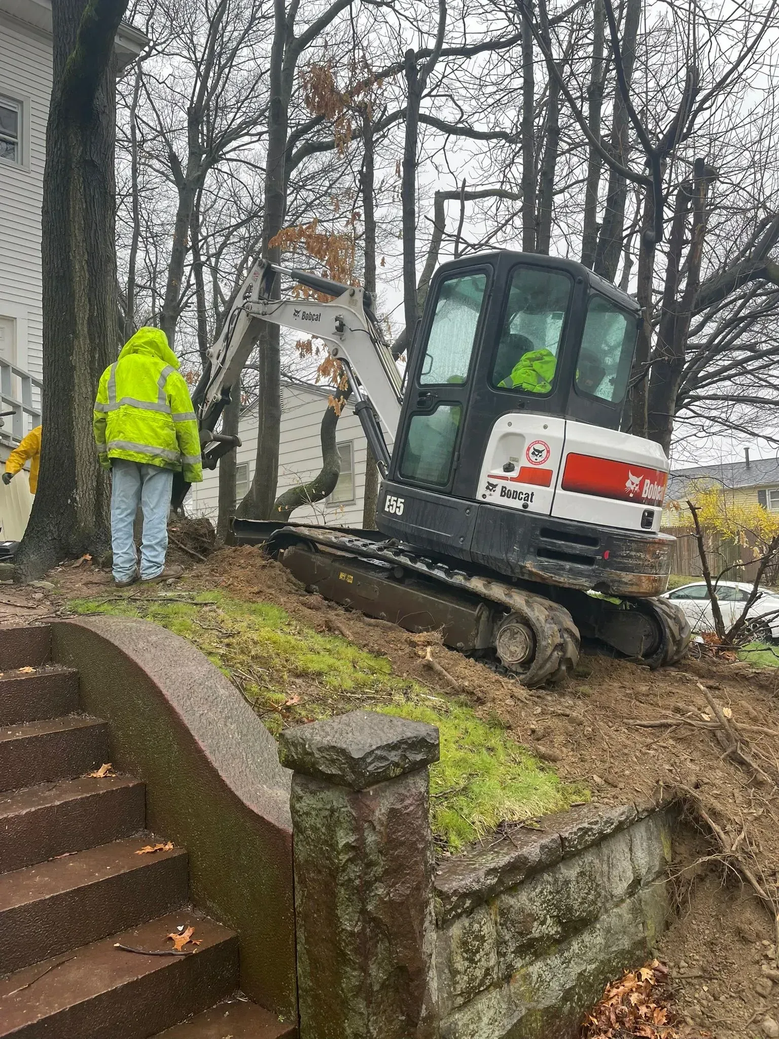 Worker beside mini excavator on a muddy slope next to a house during outdoor maintenance work