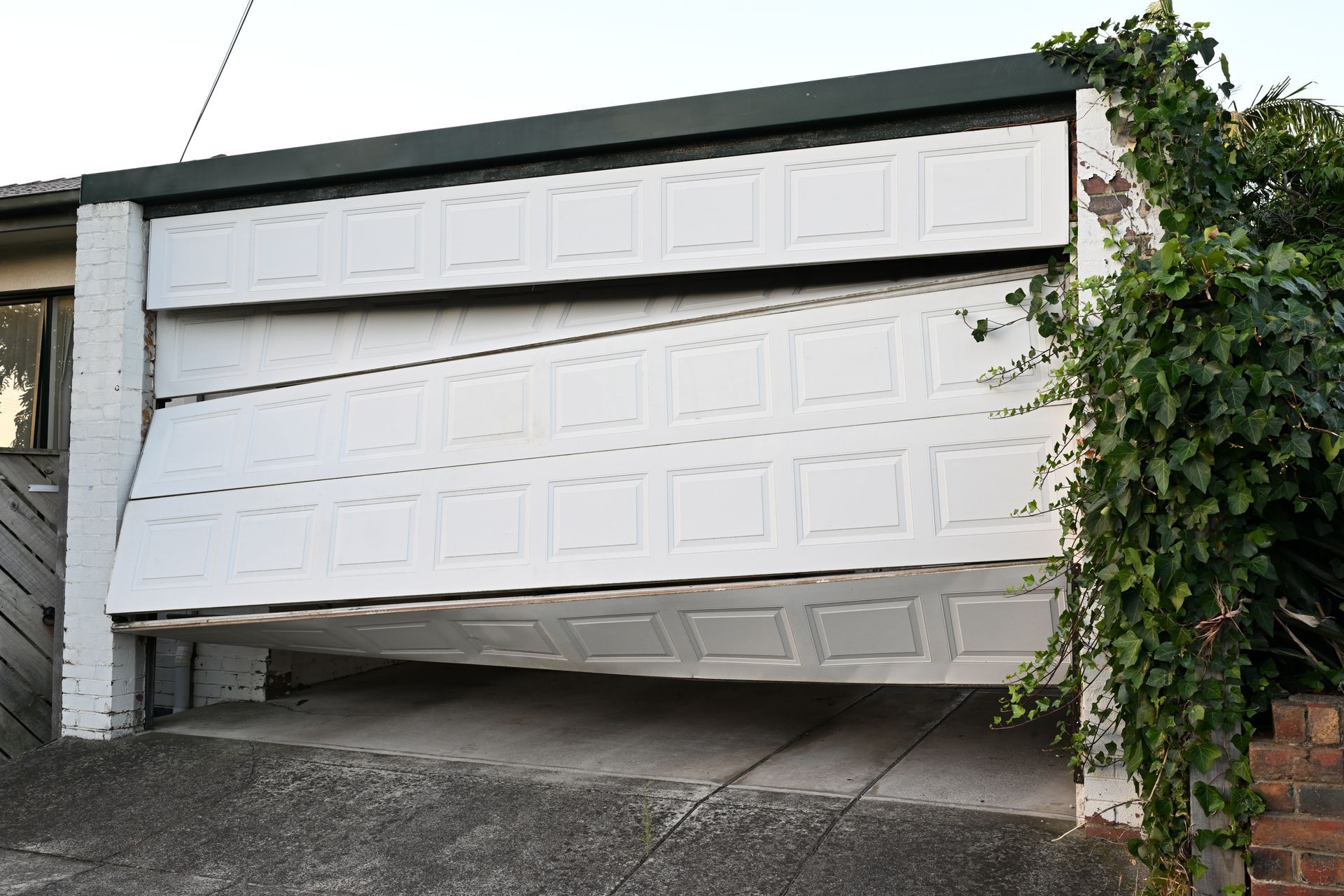 Bent, partially open garage door with damage, in front of a driveway and ivy-covered wall