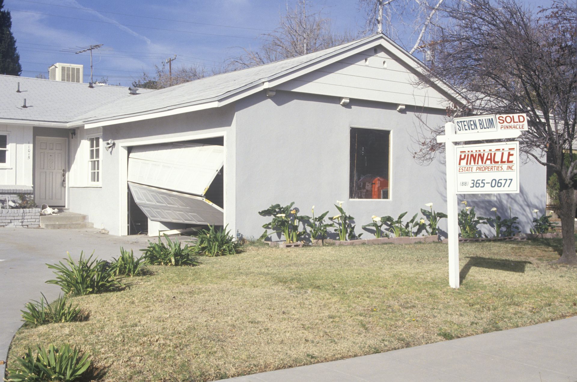 Damaged house with a collapsed garage roof and a for-sale sign in the front yard