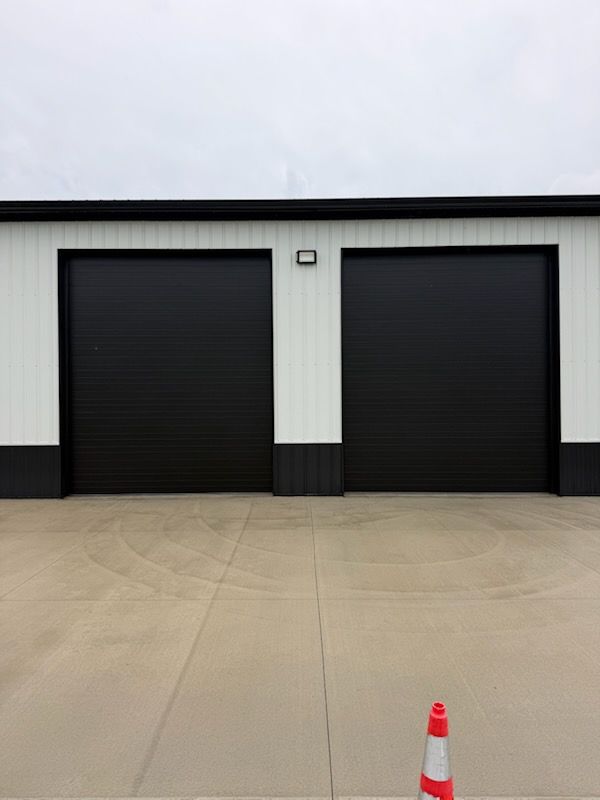Two black garage doors on a white industrial building, with a concrete lot and a traffic cone in front.