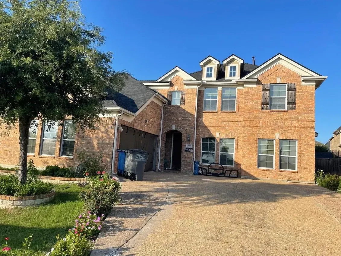 Two-story brick house with a driveway. Includes a tree on the left and trash cans in the garage.