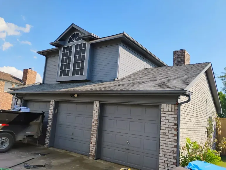 Two-story house with gray siding, brick accents, three garage doors, and a dark roof under a blue sky.