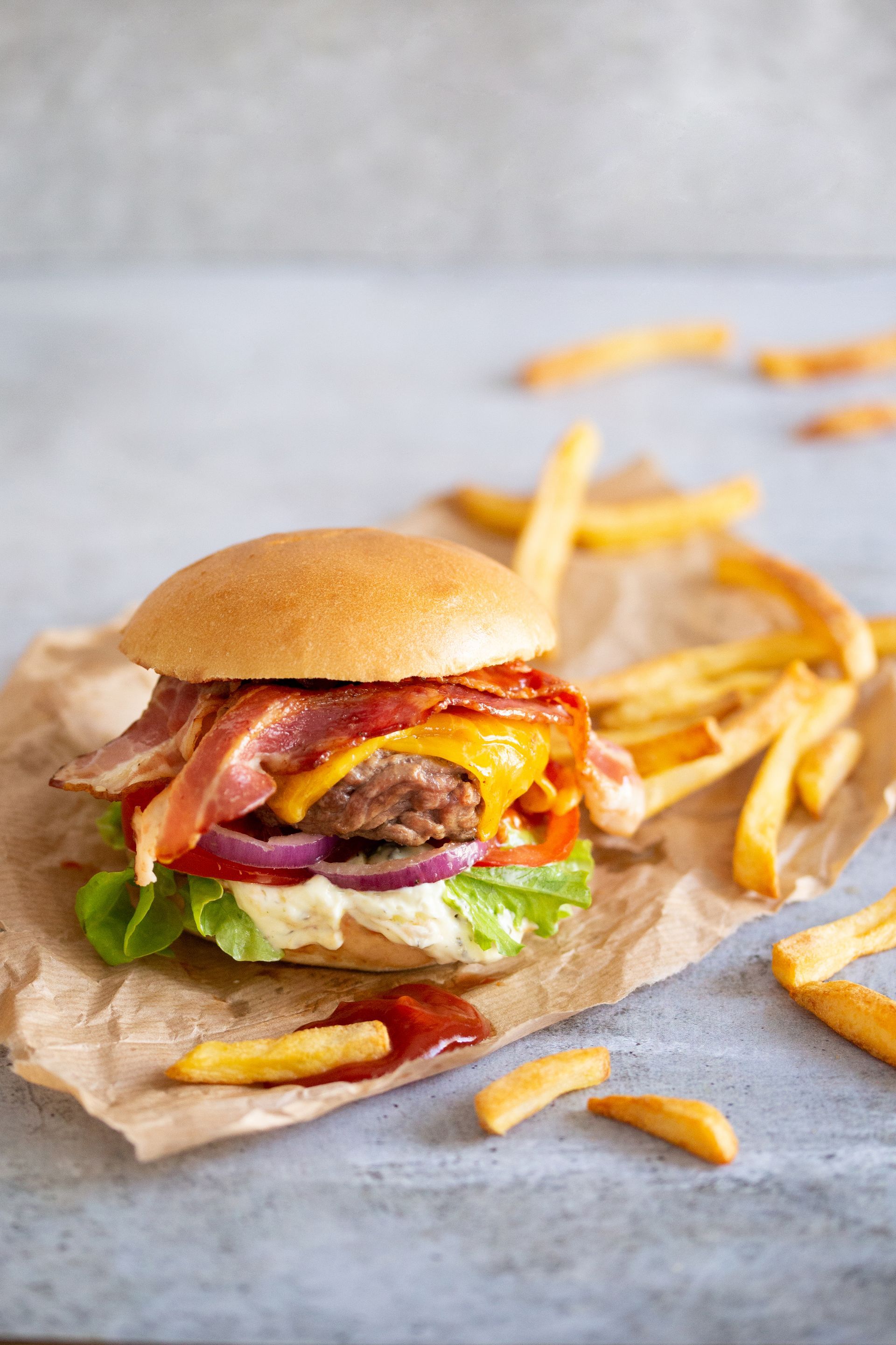 Hamburguesa con lonchas de beicon crujiente, lechuga, tomate, carne picada, salsa fundida y queso cheddar fundido, servida con patatas fritas y ketchup.