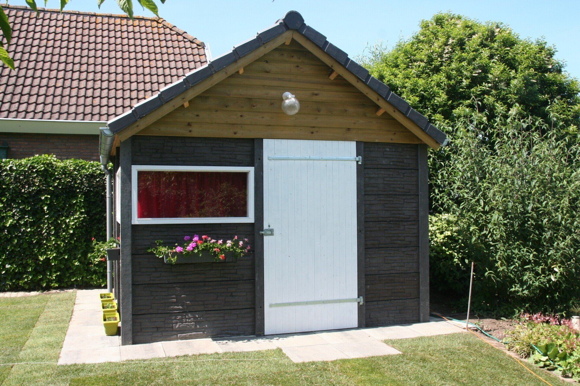 Kleine houten berging met rood dak en wit deurpaneel, uitgevoerd door A&B Betonbouw.