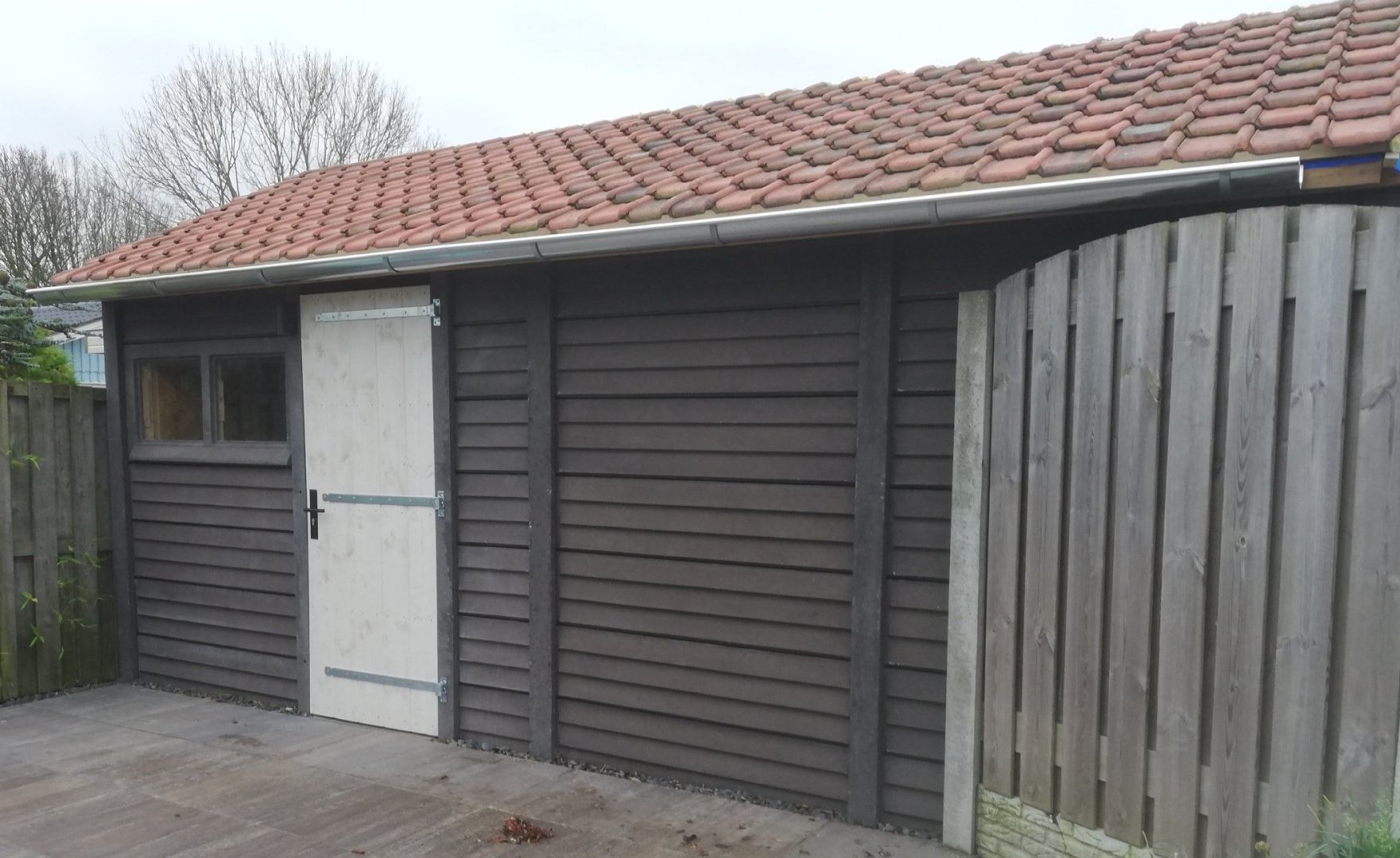 Donkere houten schuur met rood pannendak en witte deur naast een houten schutting.