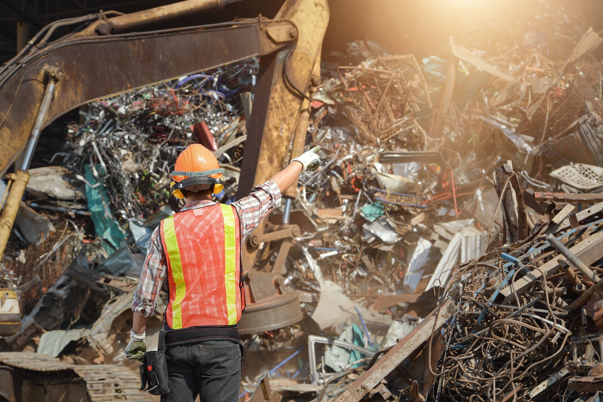 Worker in hard hat and safety vest pointing at a large pile of scrap metal at a recycling yard