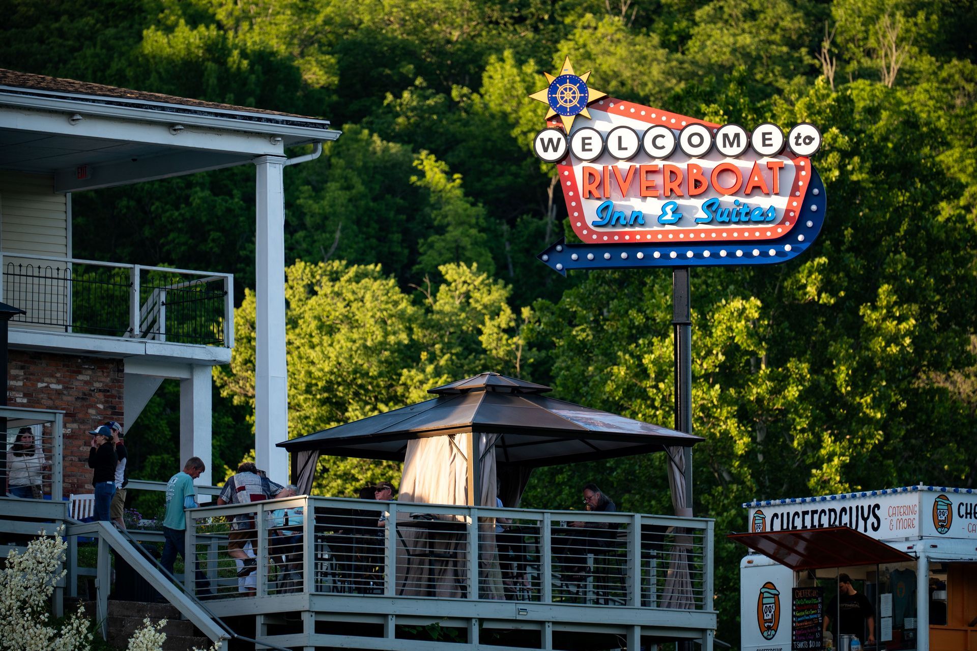 Outdoor hotel entrance with a colorful neon sign above a deck and trees in the background