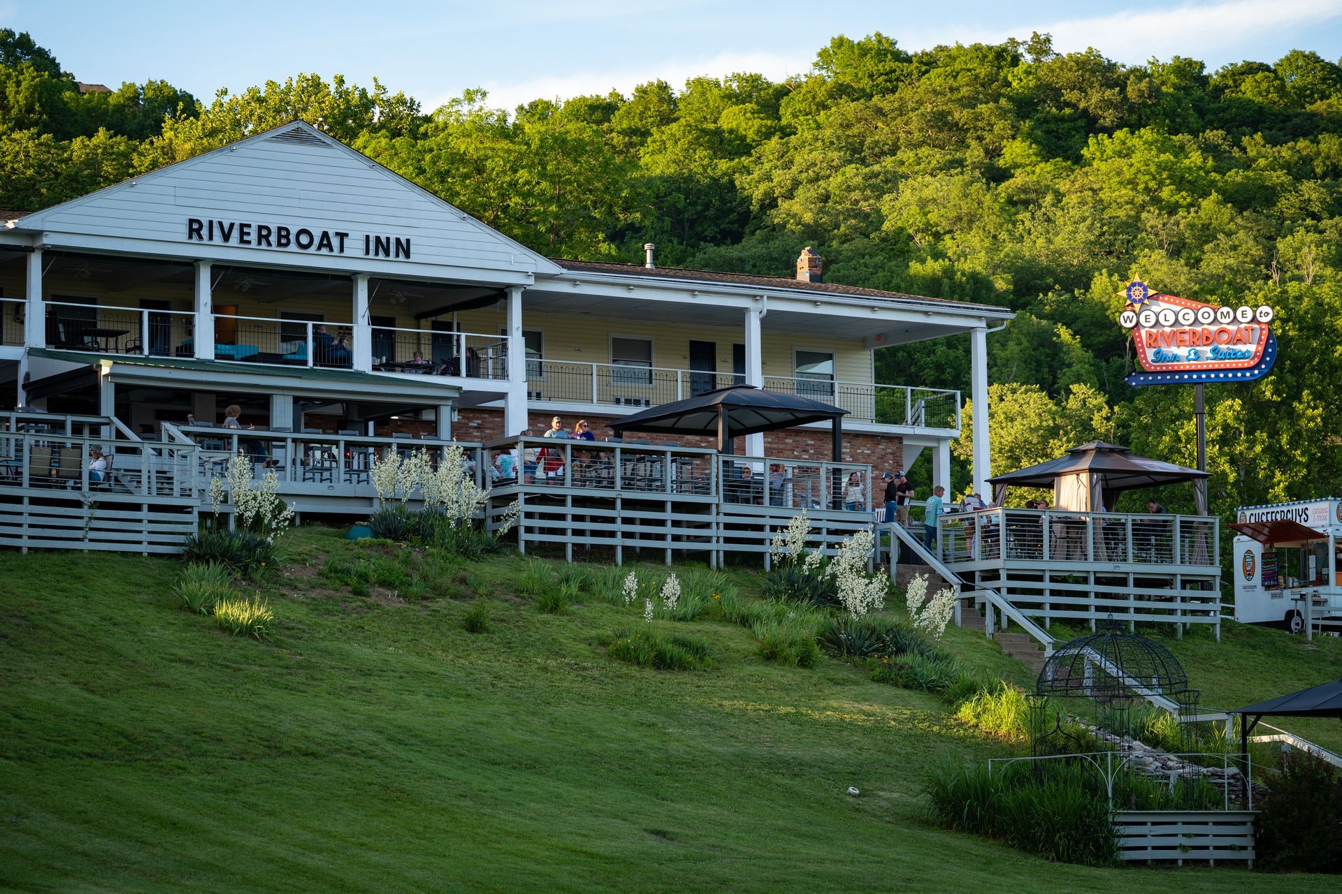 Riverside inn with white porch overlooking a grassy slope and trees, with a neon sign at right.