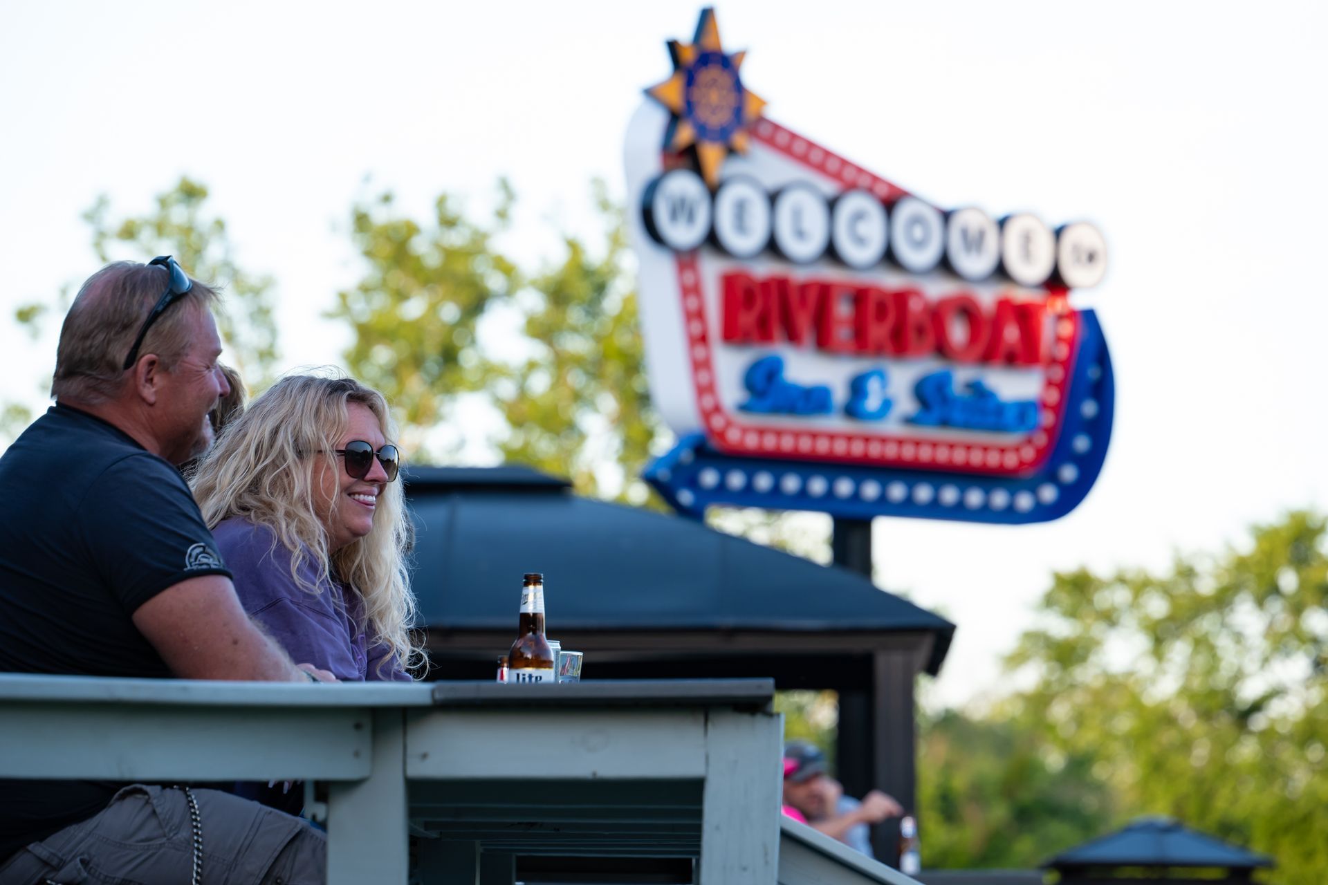 Two people sit at a table in front of a bright neon sign reading “Riverboat Inn & Suites.”