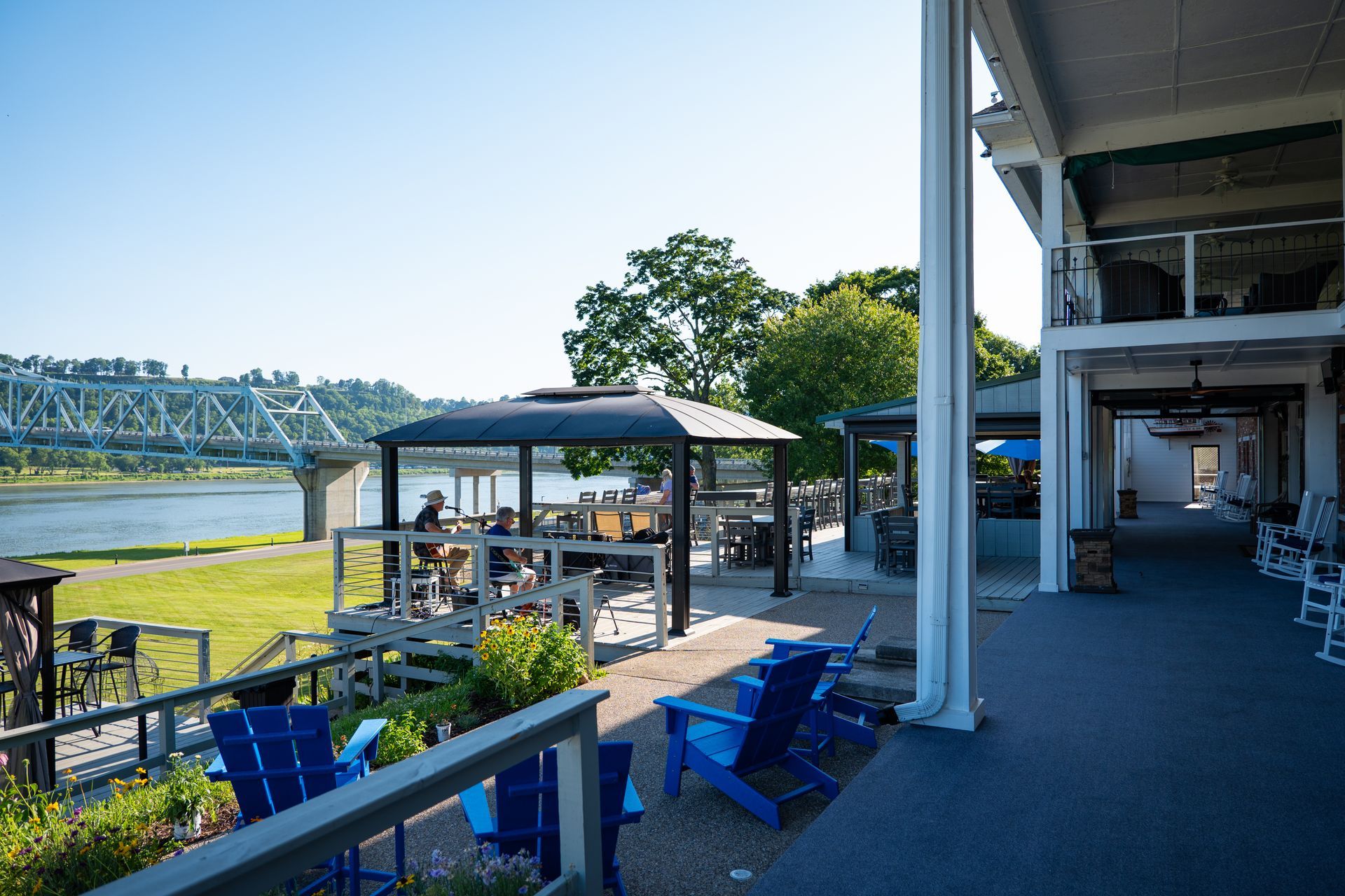 Riverside patio with blue chairs, shaded tables, and a bridge visible across the water.