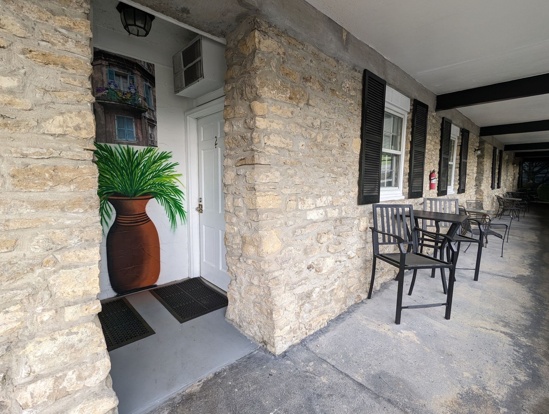 Stone porch with black metal chairs and tables beside a white doorway and potted plant