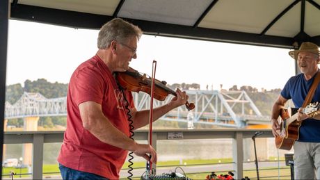 Two musicians play violin and guitar under a pavilion with a bridge in the background.
