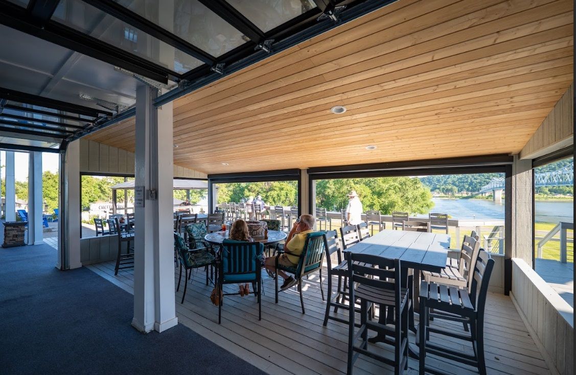 Covered outdoor patio with tables and chairs overlooking a sunny lawn and river