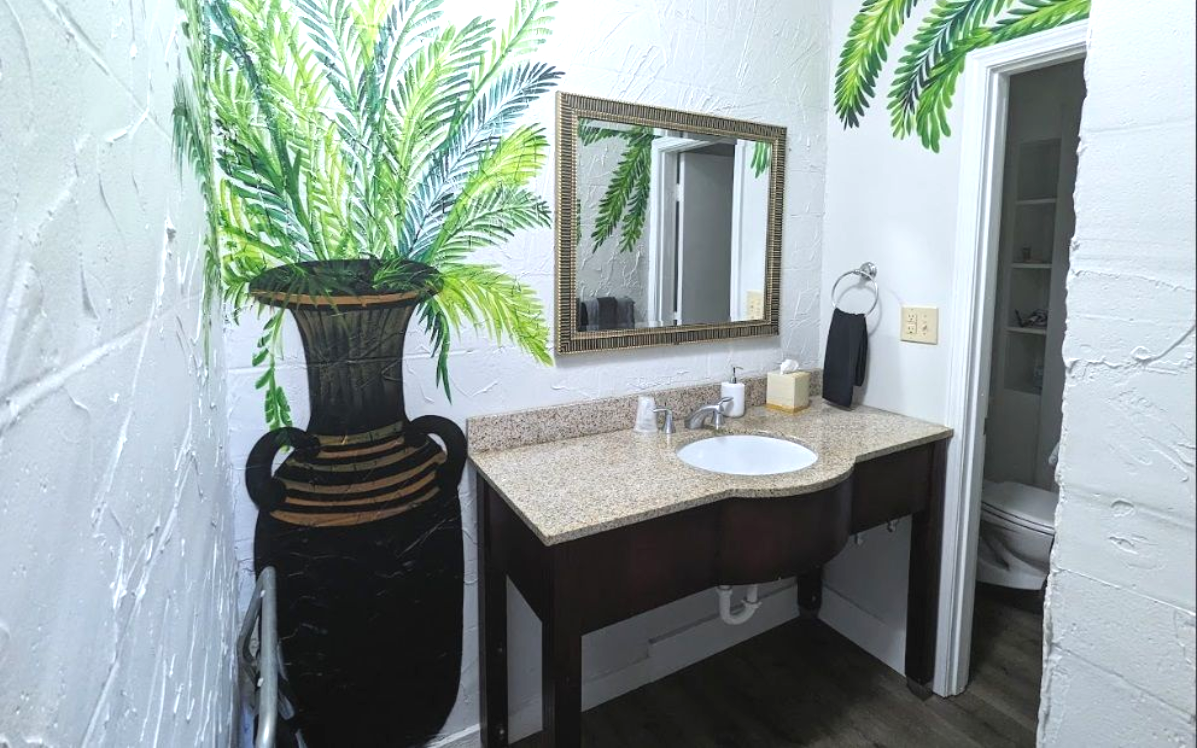 Hotel bathroom vanity with dark wood cabinet, granite countertop, mirror, and large black vase with greenery.