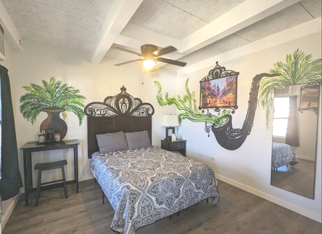 Hotel room with ornate black bed, patterned bedding, ceiling fan, wall mirror, and potted plants on stands