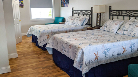 Bright hotel room with two queen beds, wooden floor, and a window with teal chair in the back.