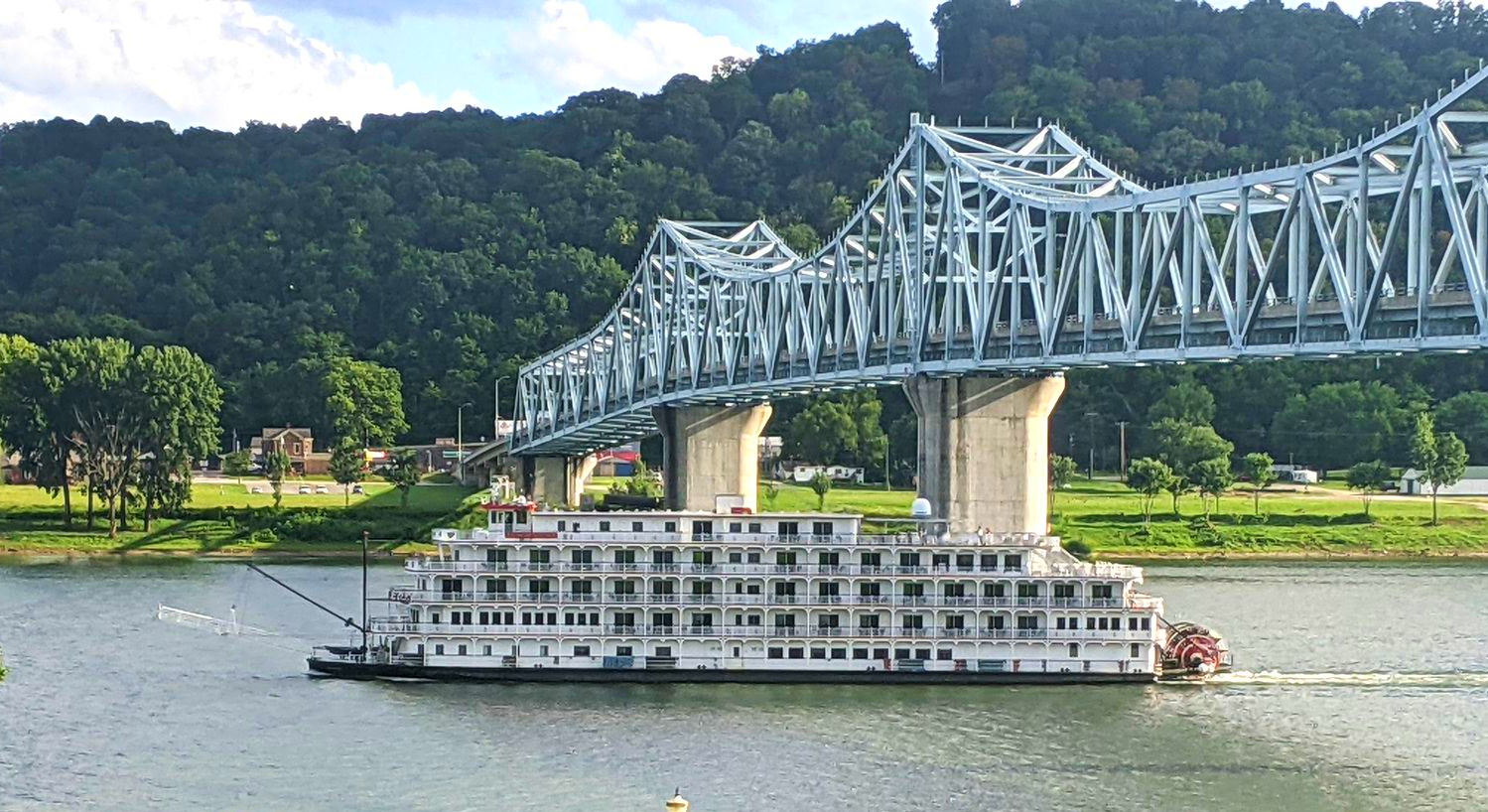 Steamboat on a river beneath a steel bridge with forested hills in the background