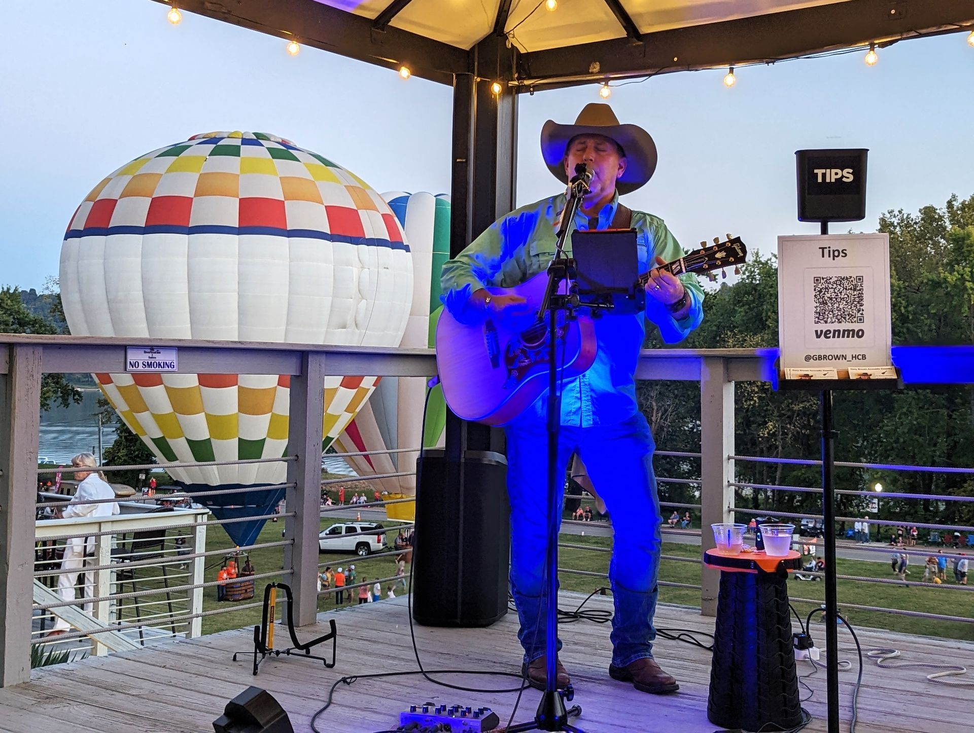 A man is playing a guitar and singing into a microphone in front of a hot air balloon.