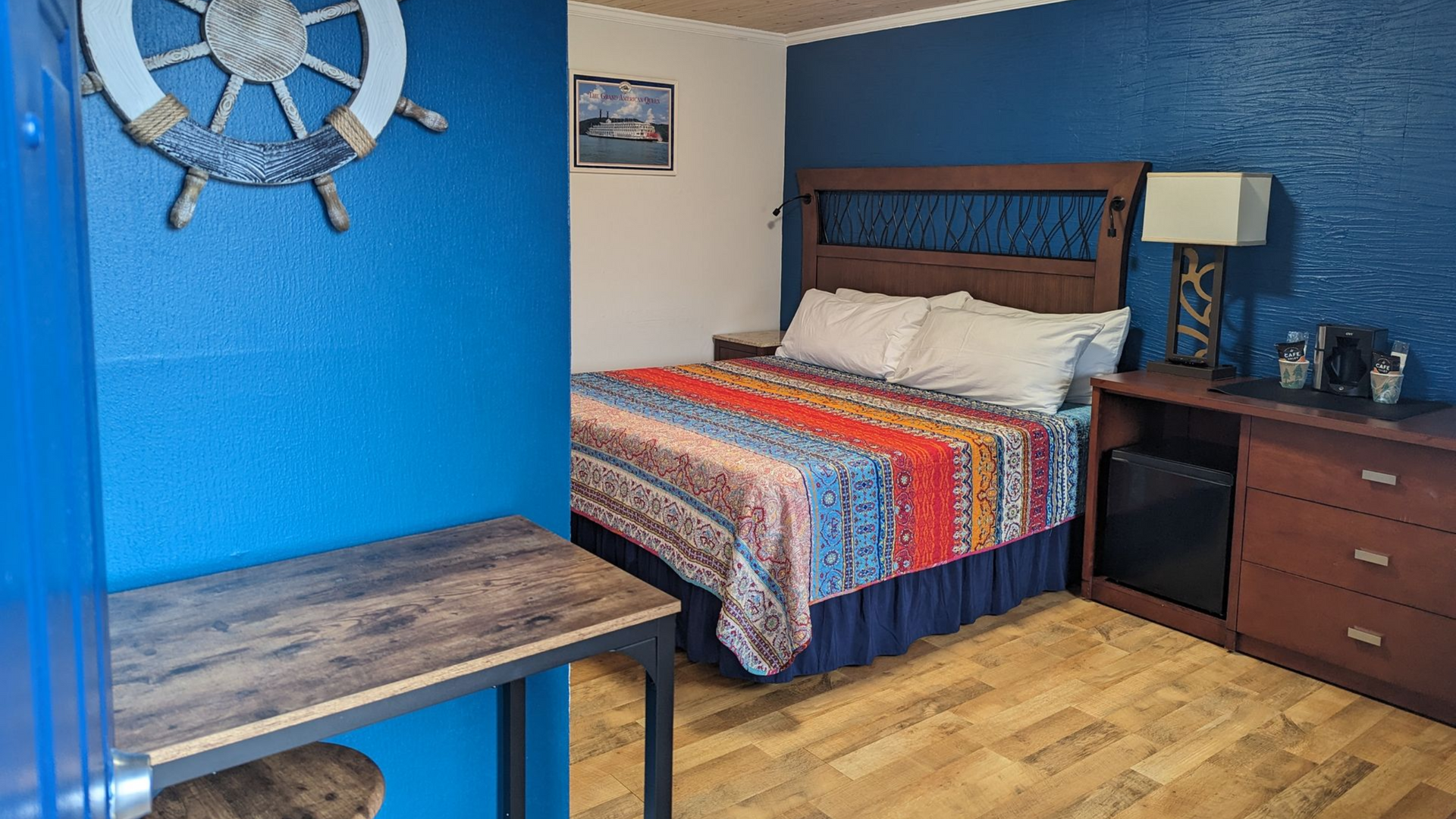 Blue-walled hotel room with a bed, wooden desk, dresser, lamp, and ship wheel wall decor.