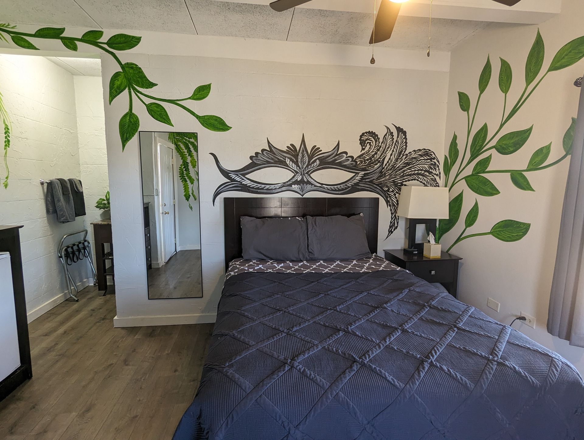 Bedroom with gray bedspread, decorative headboard, wall mirror, and green vine mural.