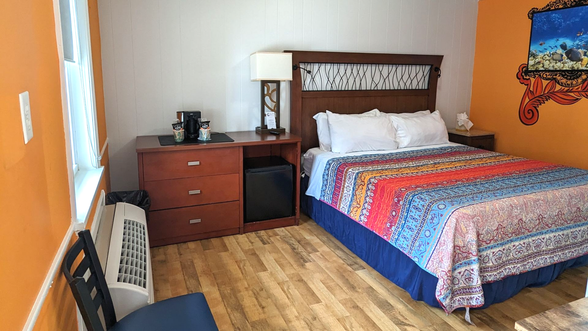 Hotel room with a bed, dresser, lamp, and wall art beside a window and radiator.