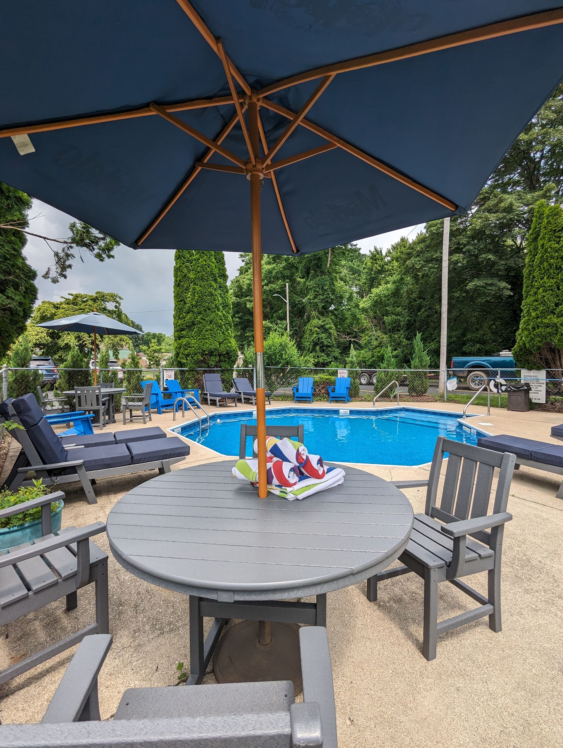 Poolside table and chairs under a large blue umbrella, overlooking a swimming pool and lounge chairs.