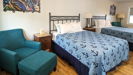 Cozy hotel room with two beds, teal armchair, wooden nightstands, and nautical-themed bedding.