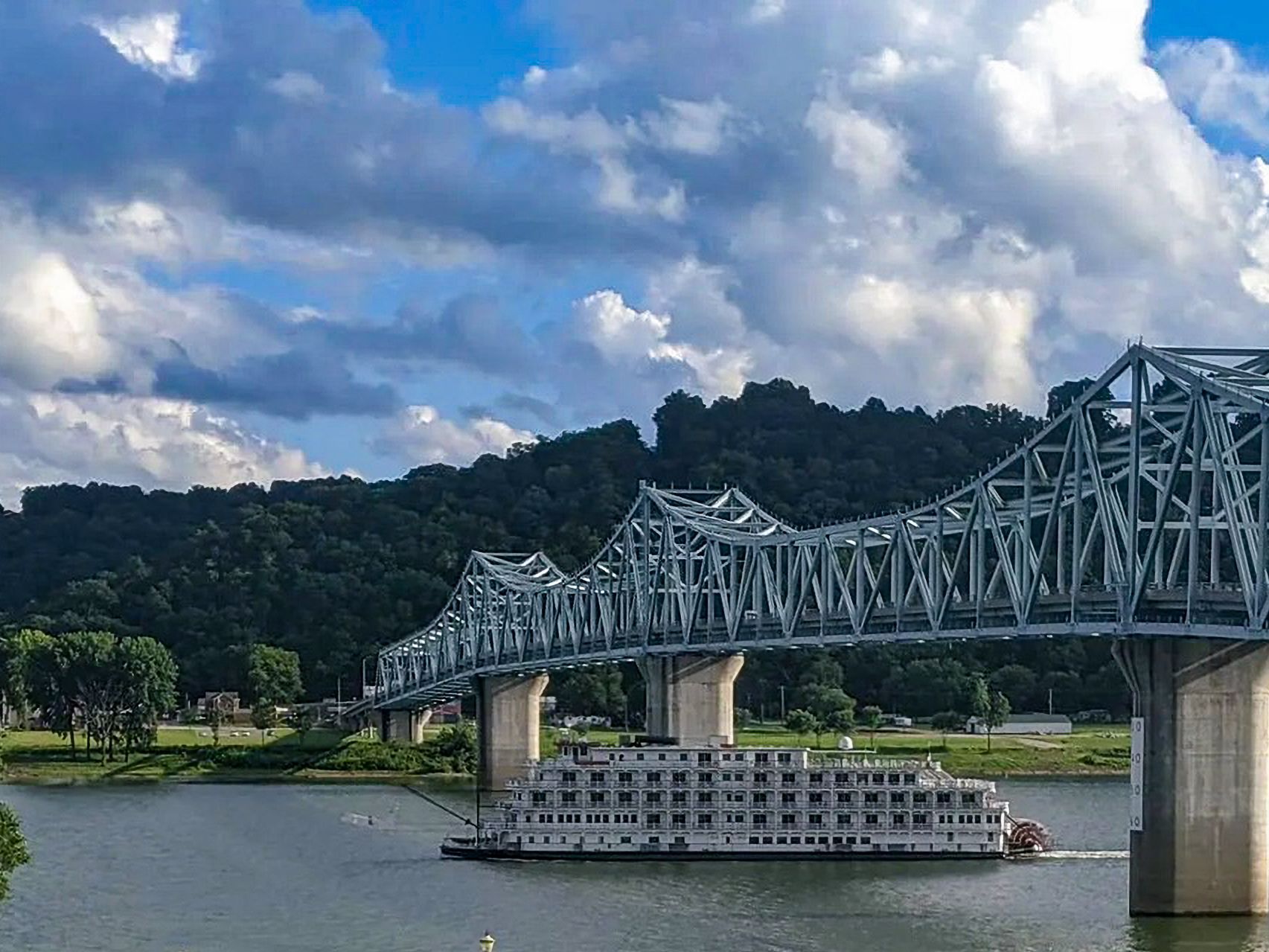 Steel truss bridge over a river with a white riverboat beneath and green hills under cloudy skies