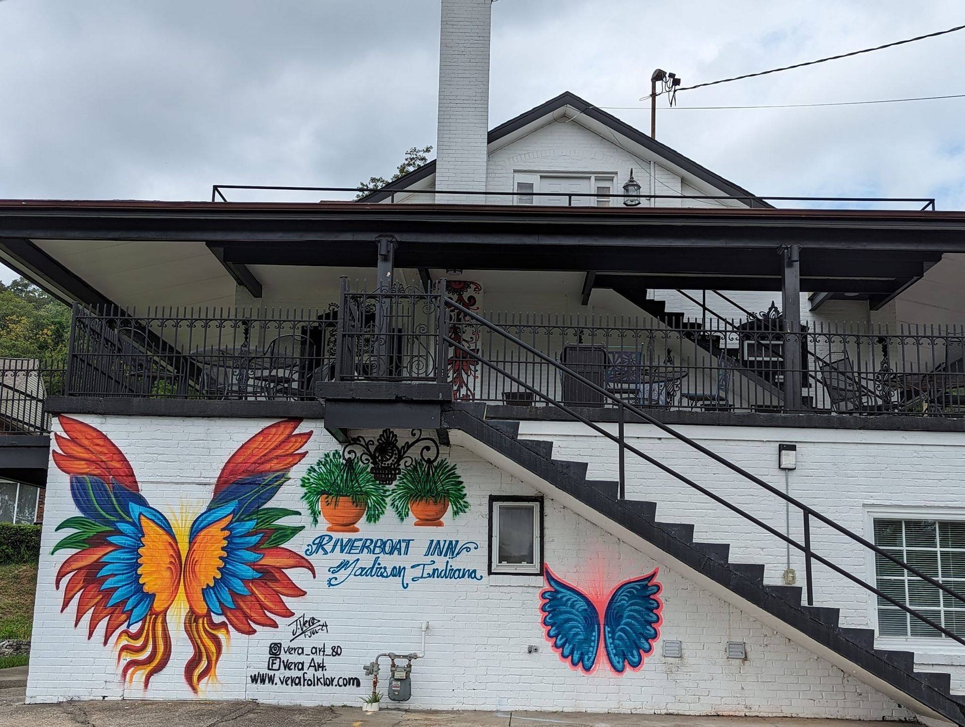 Mural of a colorful butterfly on a white brick wall beneath a building porch and stairs