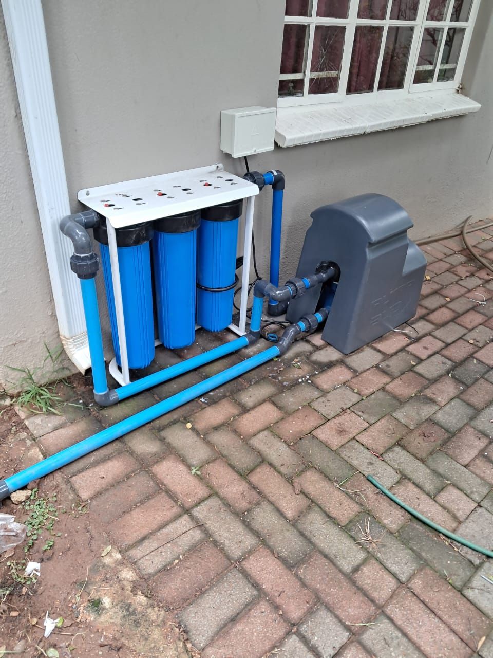 A water filter is sitting on a brick sidewalk next to a window.