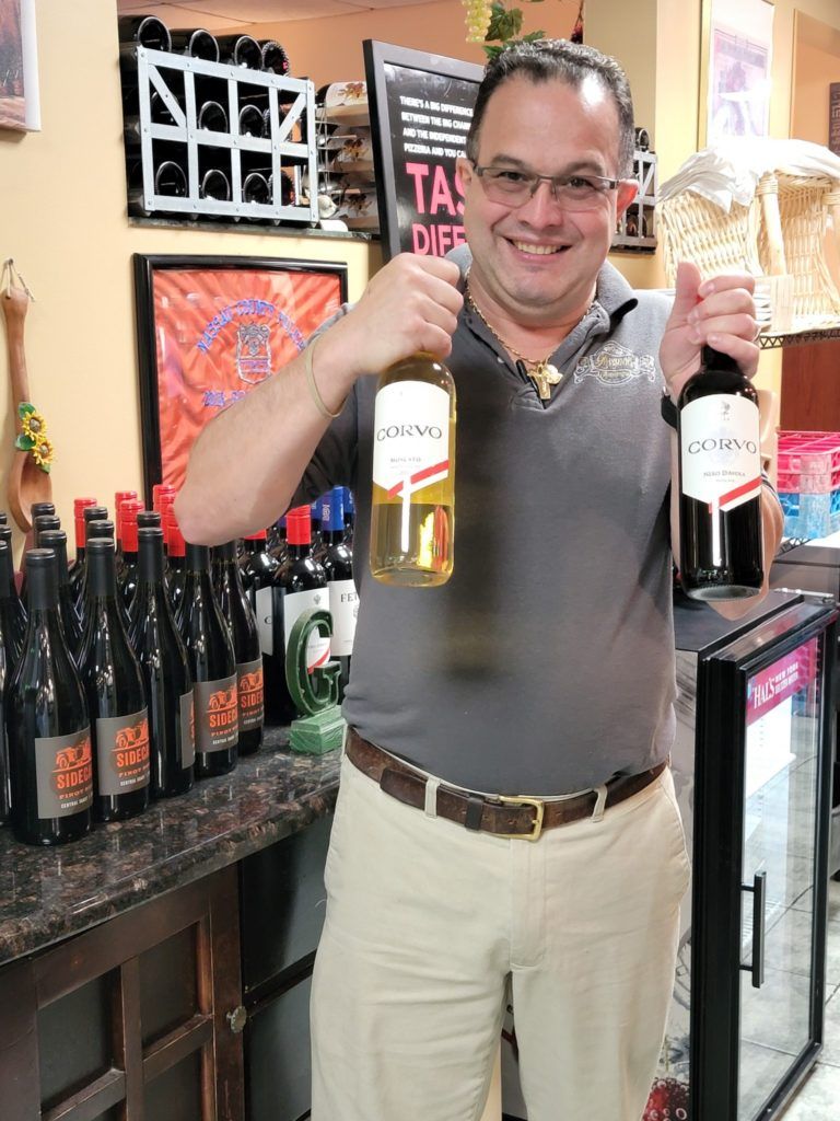 Smiling man holding two wine bottles in a liquor store, with shelves of wine behind him