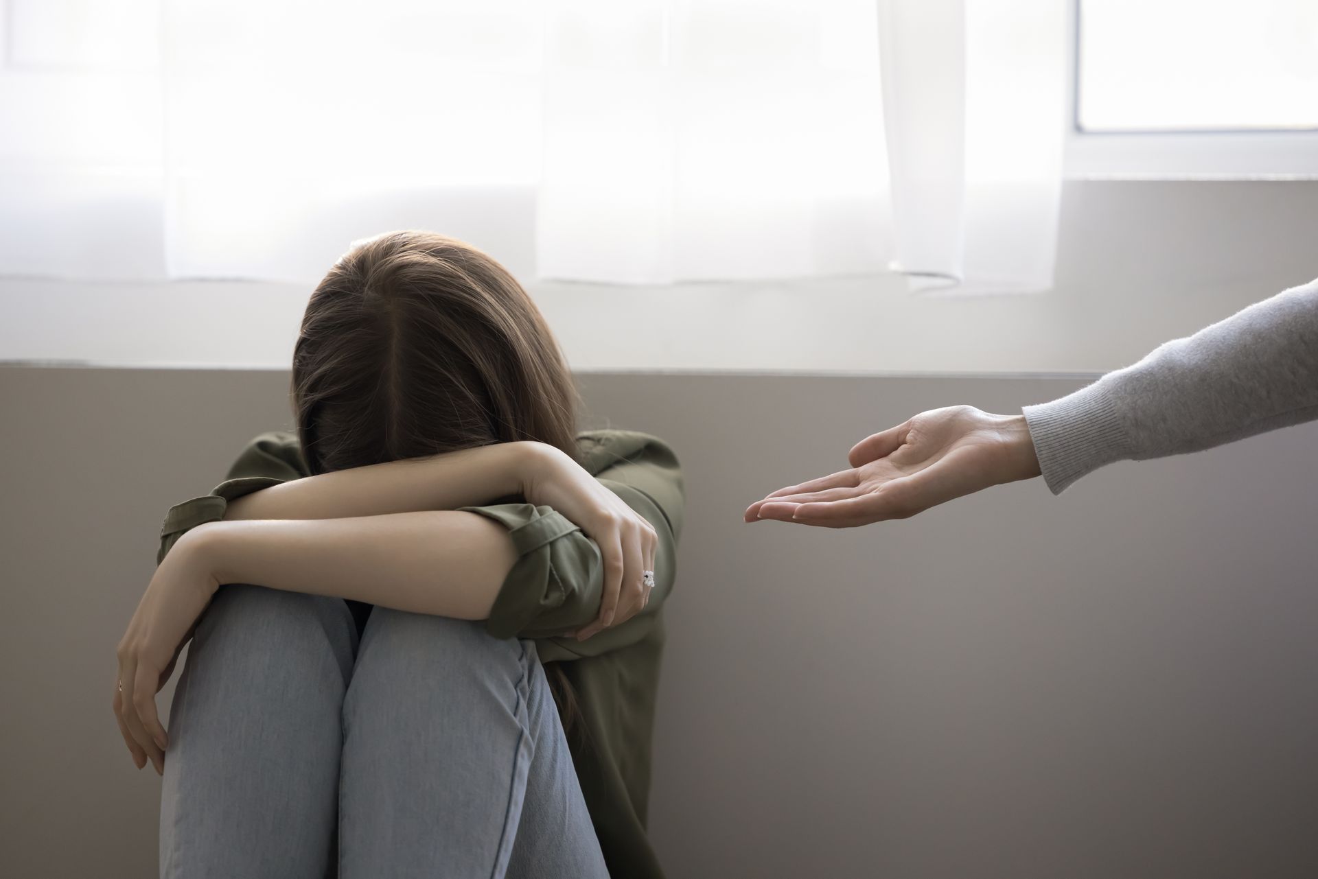 Person sitting on the floor, curled up and crying, while another hand reaches out in comfort.