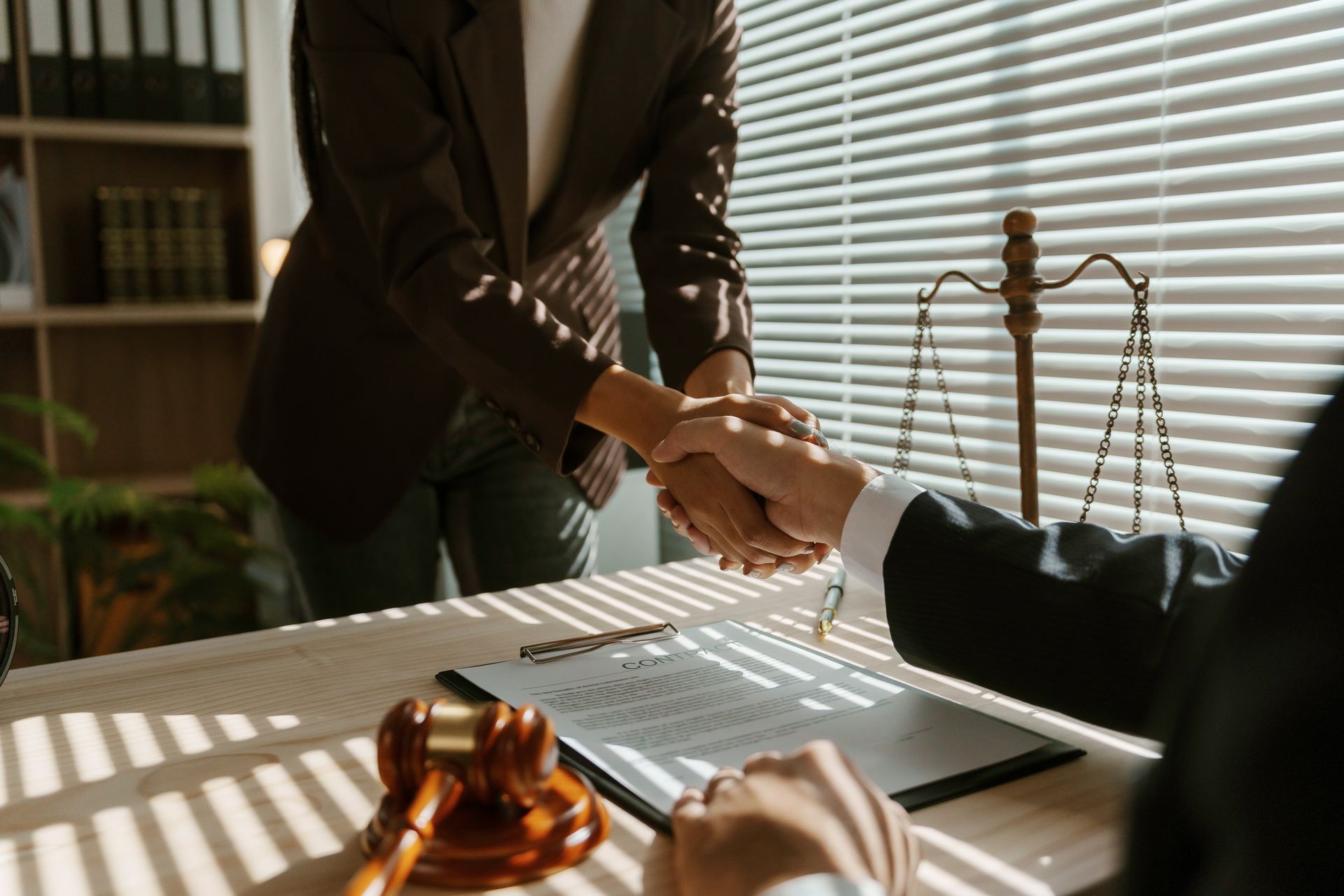 Lawyers shaking hands over a desk with a gavel and scales of justice in a sunlit office