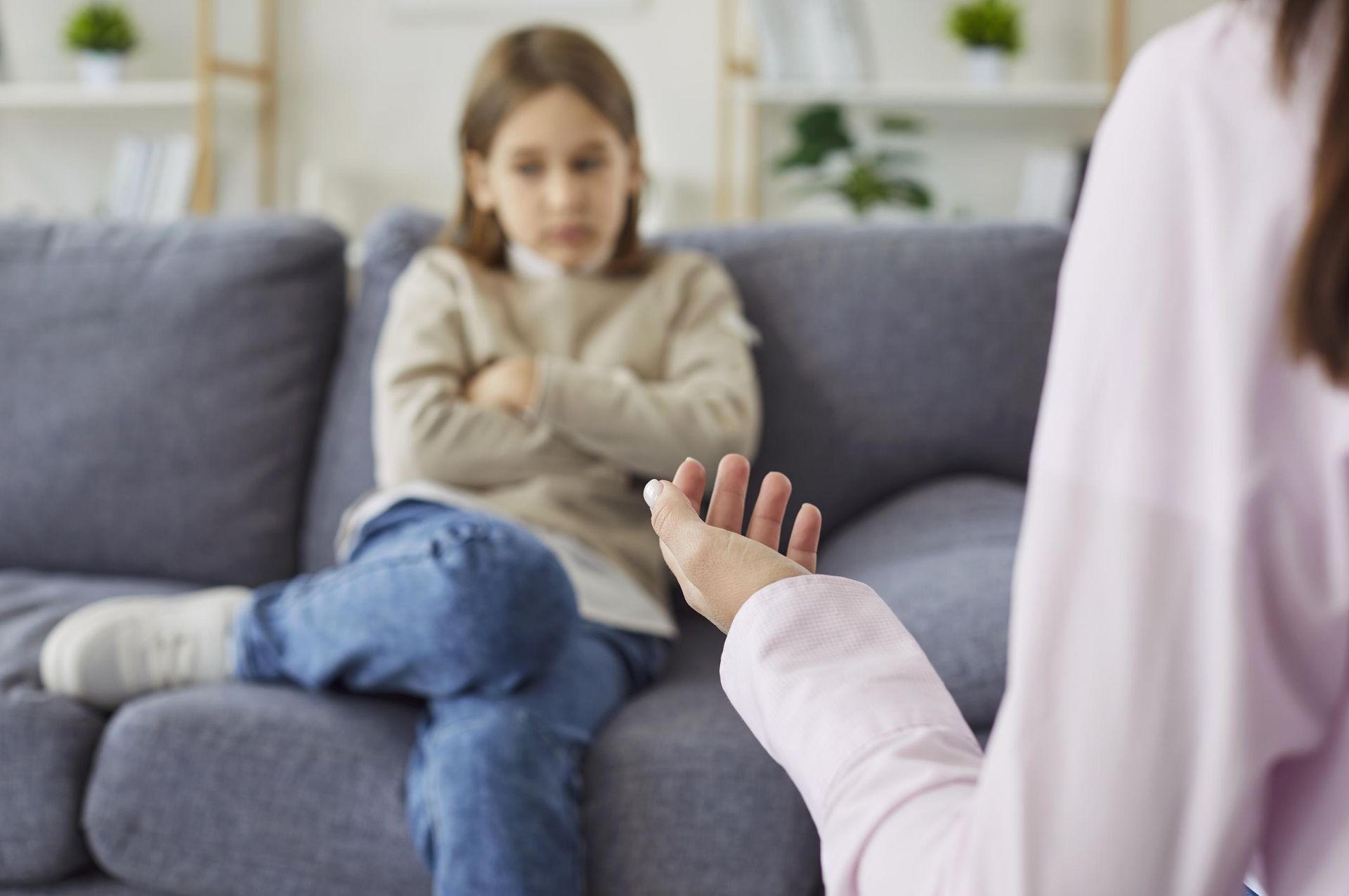 Child sitting on a couch, arms crossed, while an adult gestures toward them in a living room.