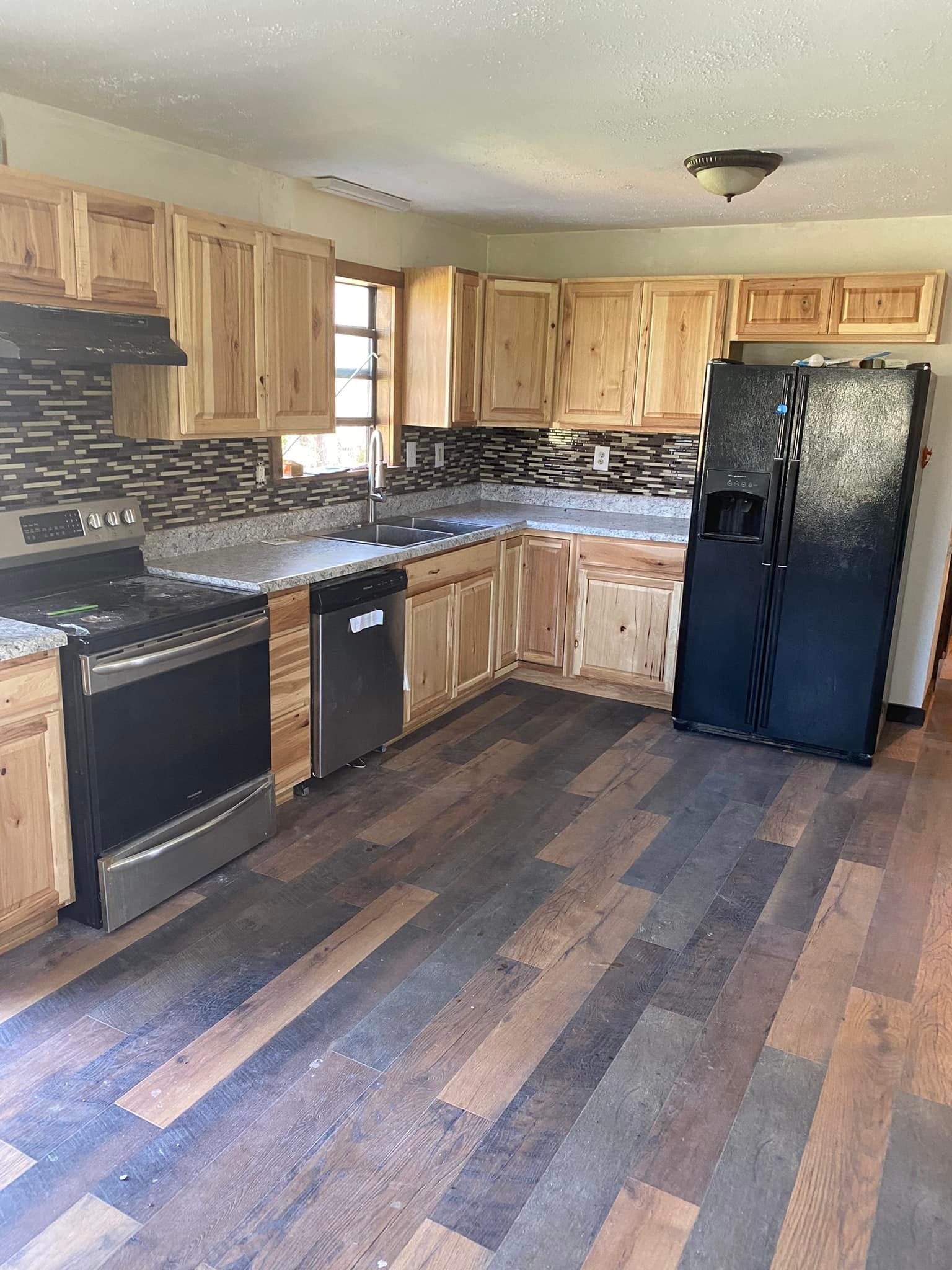 A kitchen with light wood cabinets, dark countertops, a tile backsplash, stainless steel oven, and dark plank flooring.