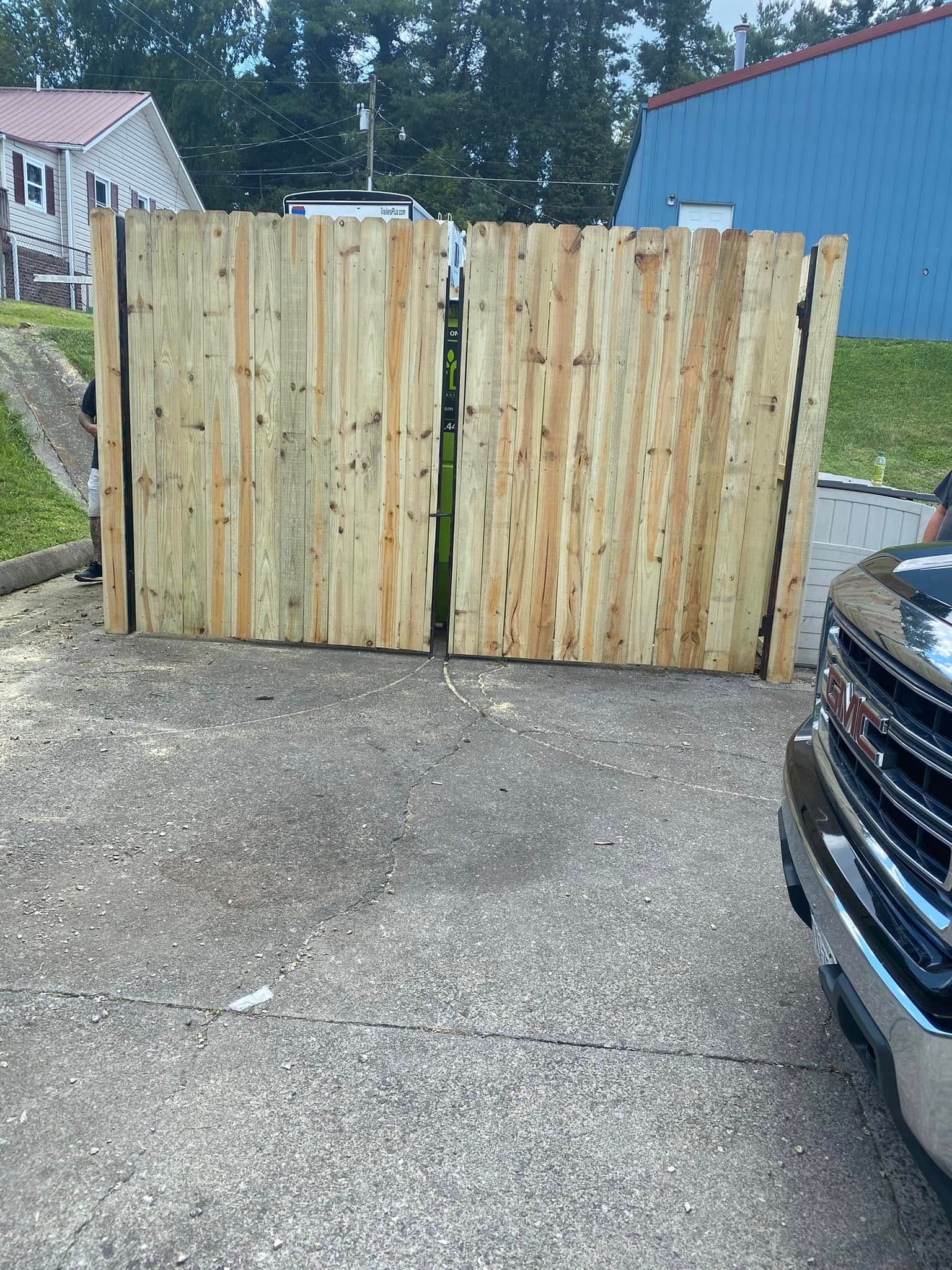 Two side-by-side wooden fence panels stand on a paved driveway next to the front of a dark vehicle.