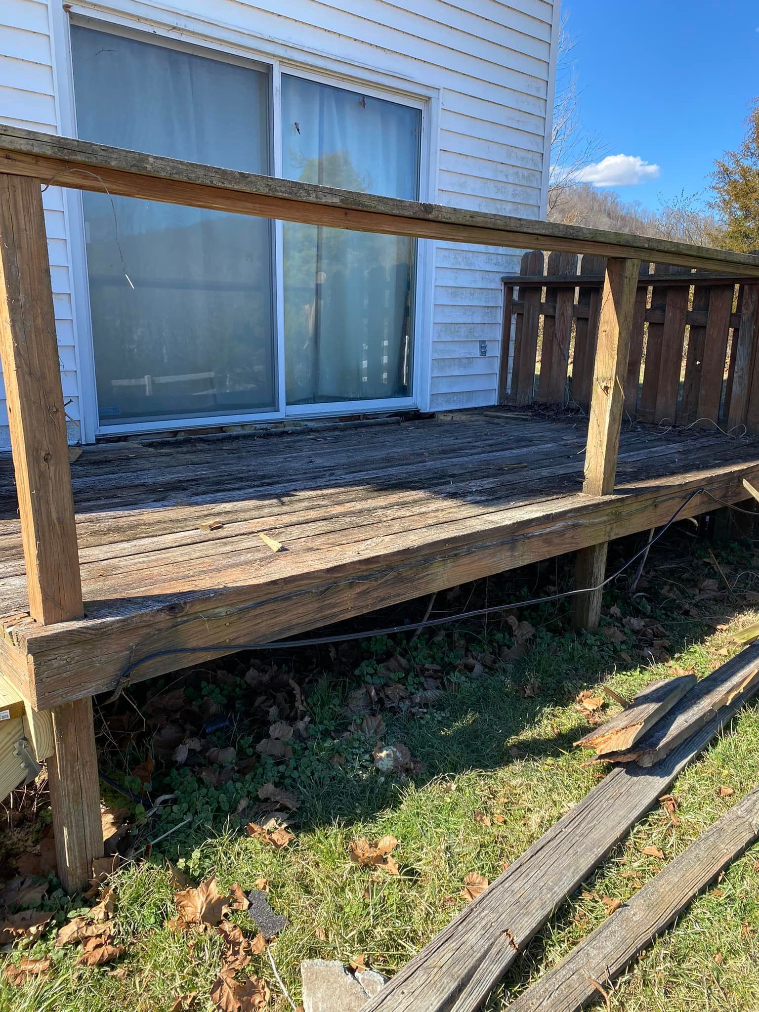 A weathered, wooden deck with a simple railing stands outside the sliding glass door of a white, vinyl-sided home.