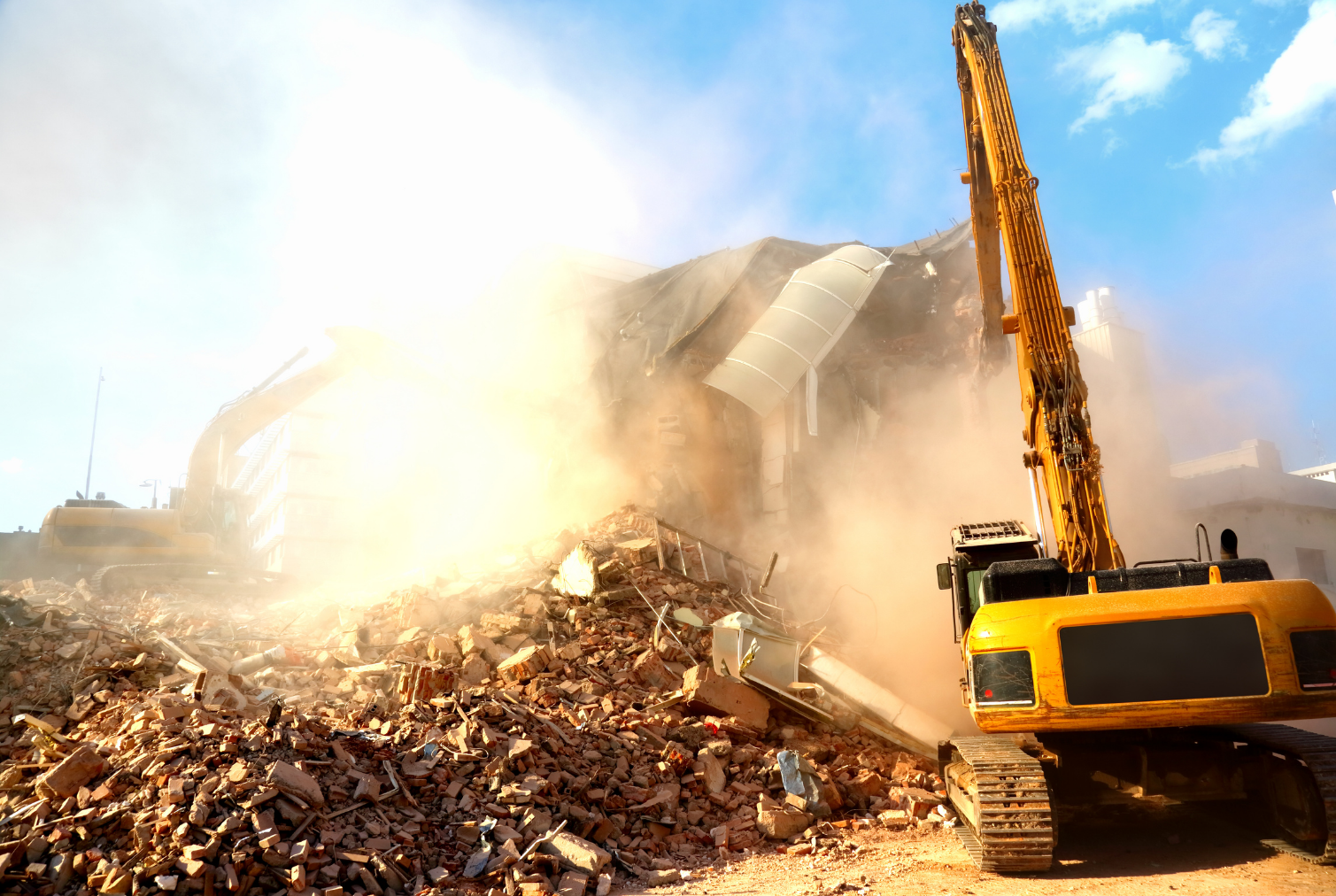 Yellow construction excavators demolish a building, kicking up a large cloud of dust over a pile of debris.