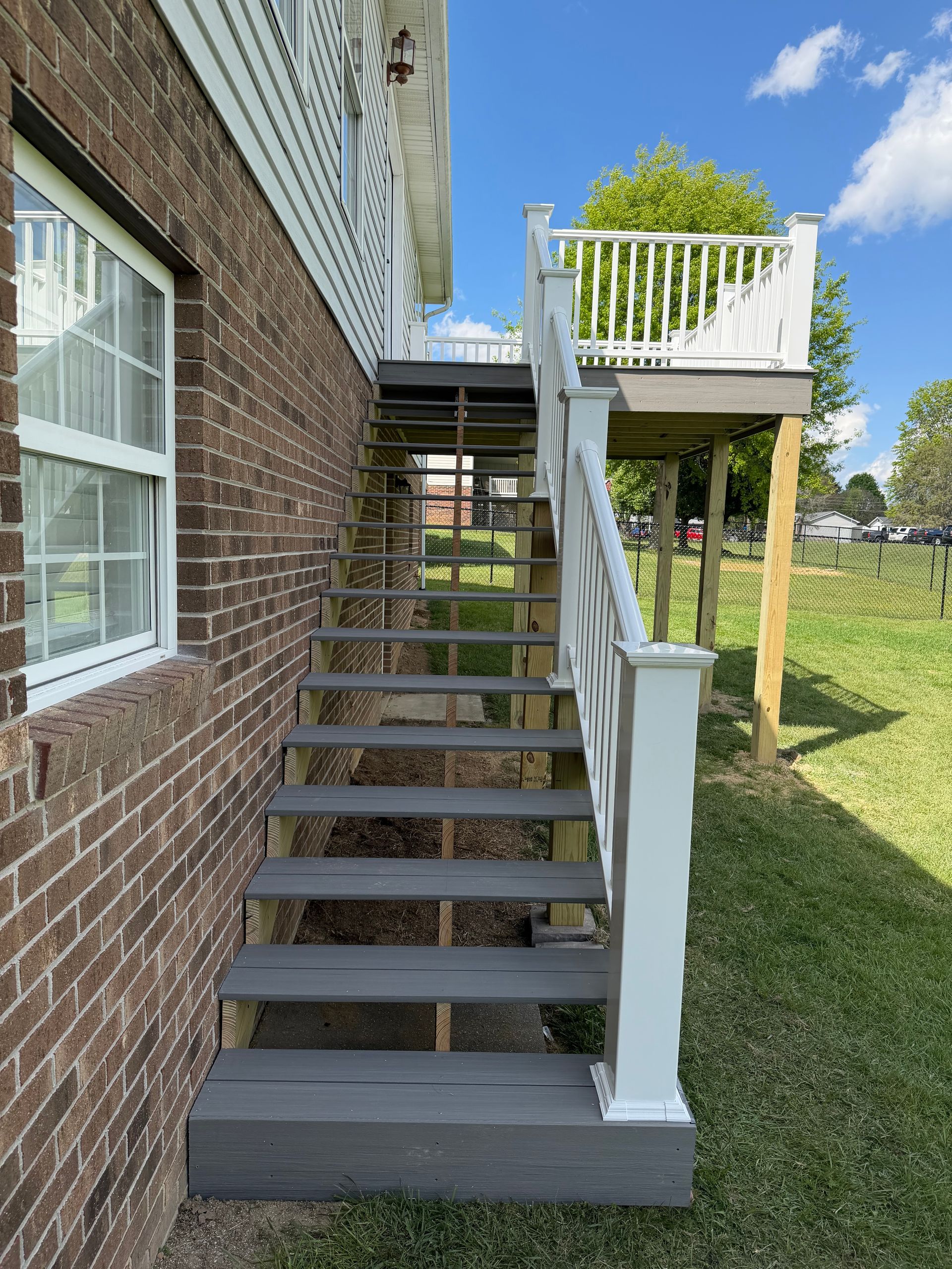 Outdoor wooden stairs beside a brick house lead to a white-railed deck over a grassy yard.