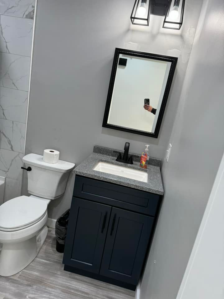 A modern bathroom with a navy vanity, grey countertop, rectangular mirror, toilet, and contemporary light fixture.