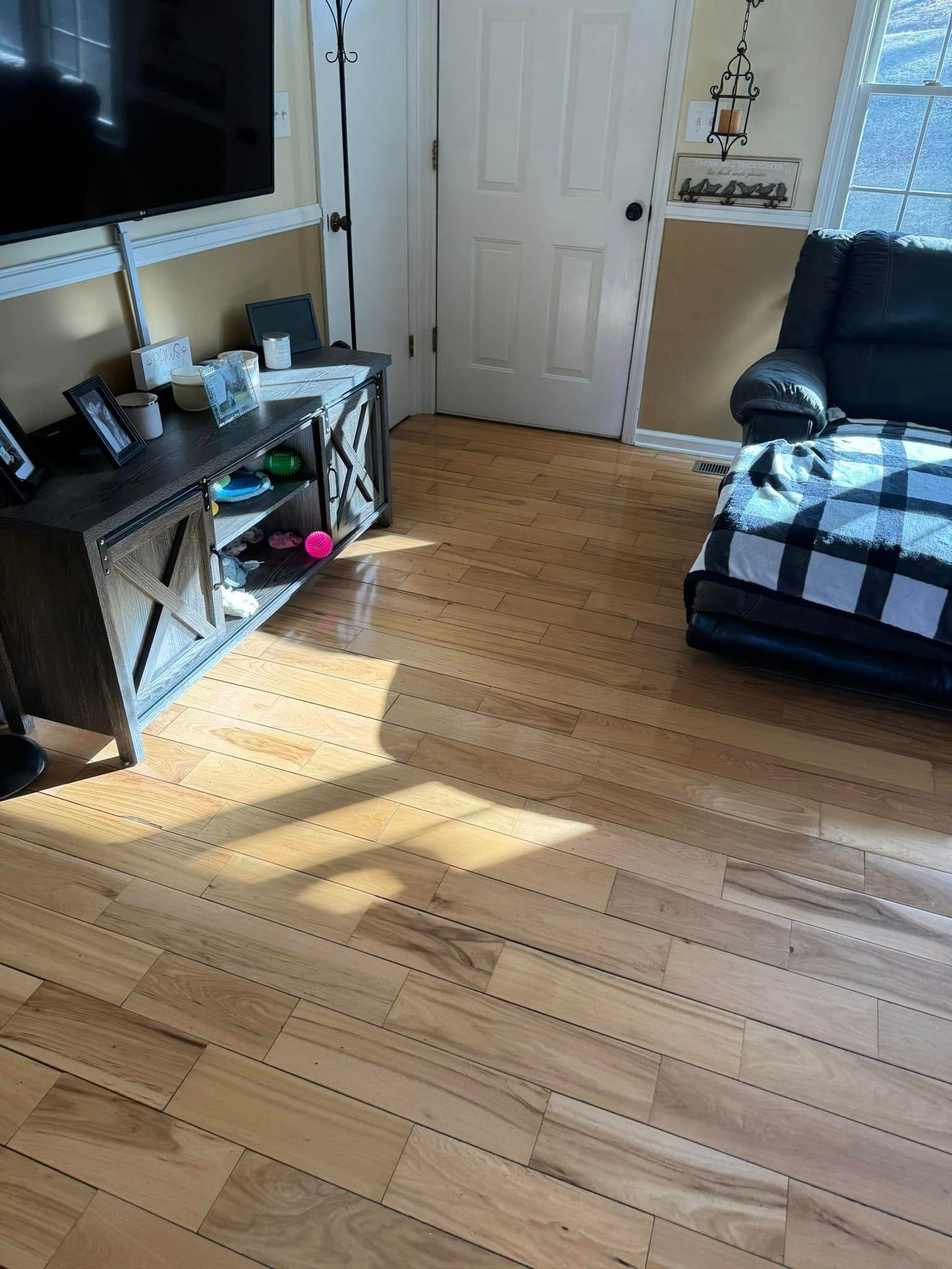 A living room view showing light wood flooring, a television on a gray media console, and a black armchair.