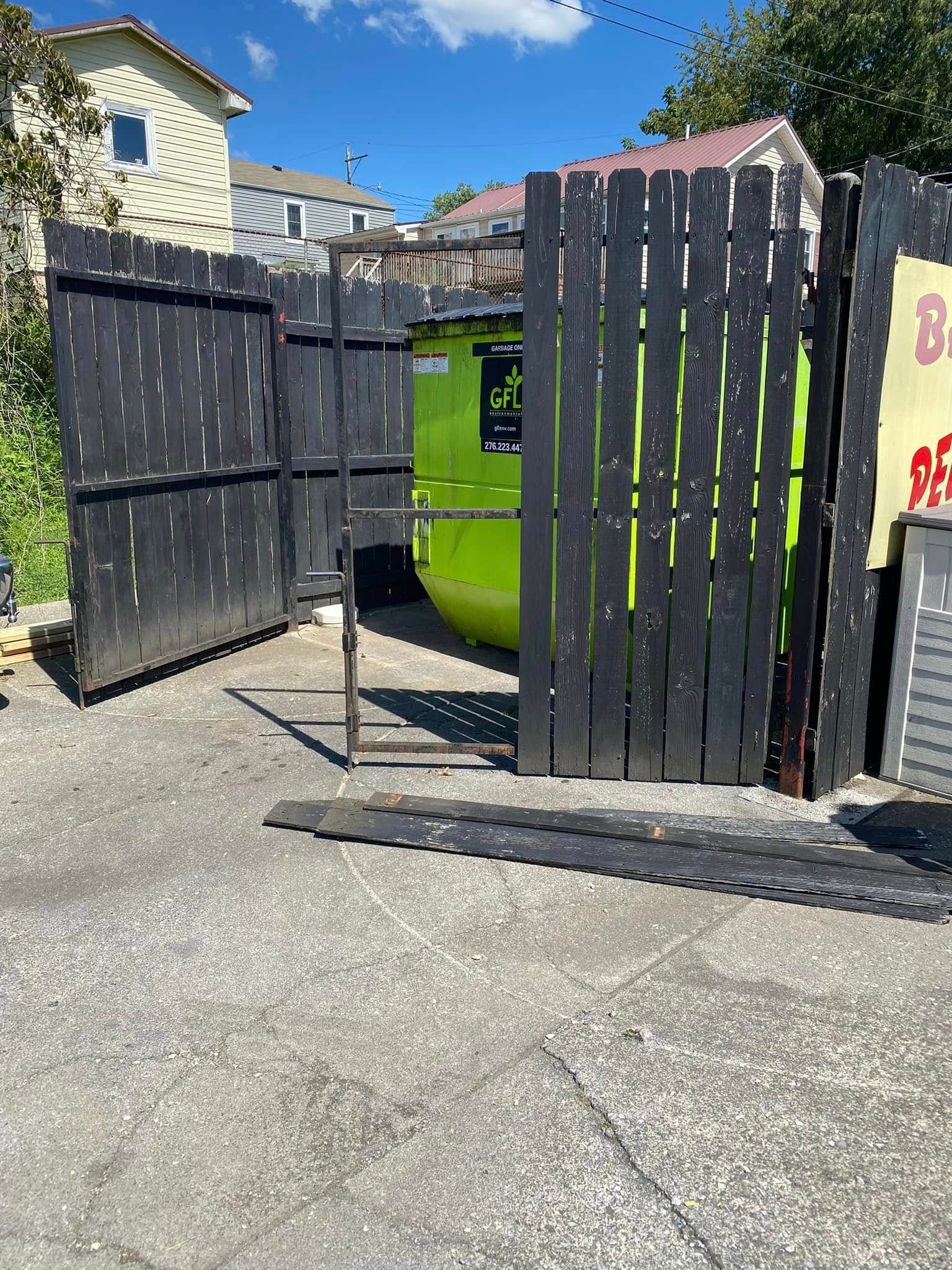 A bright green dumpster is partially enclosed by a black wooden fence in an outdoor gravel lot.
