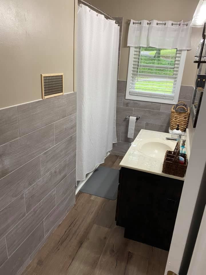 An after picture of the same bathroom featuring wood-look tile flooring, gray tile wall accents, a dark vanity,