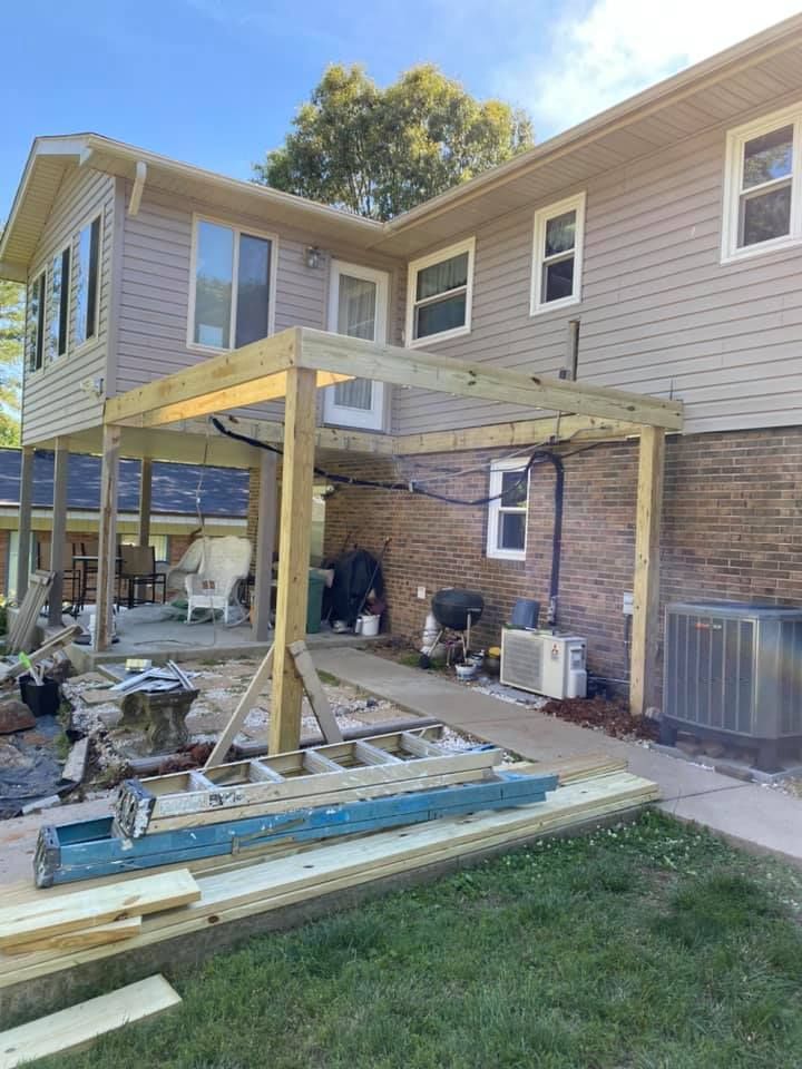 A wooden deck structure under construction attached to a two-story house with beige siding and a brick lower level.