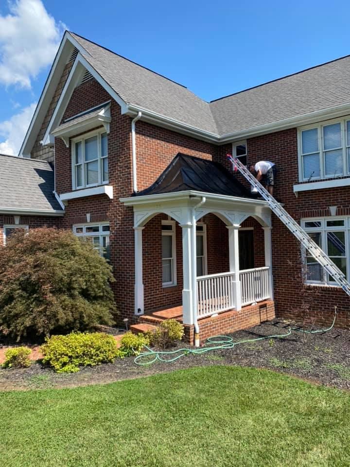 A worker on a ladder performs maintenance on the metal roof of a brick home's front porch on a sunny day.