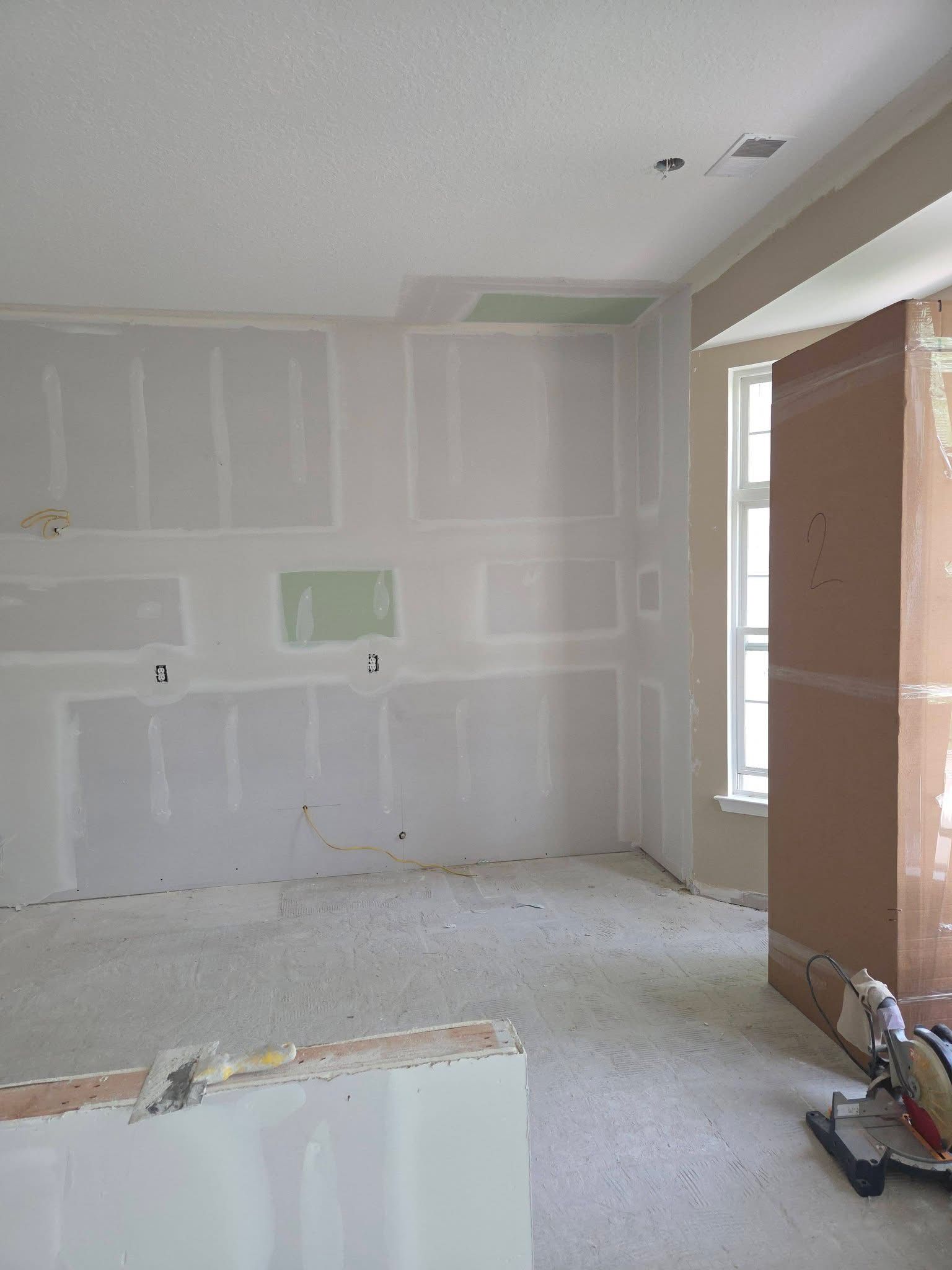 Unfinished room with drywall, white dust-covered floor, window light, and construction materials.