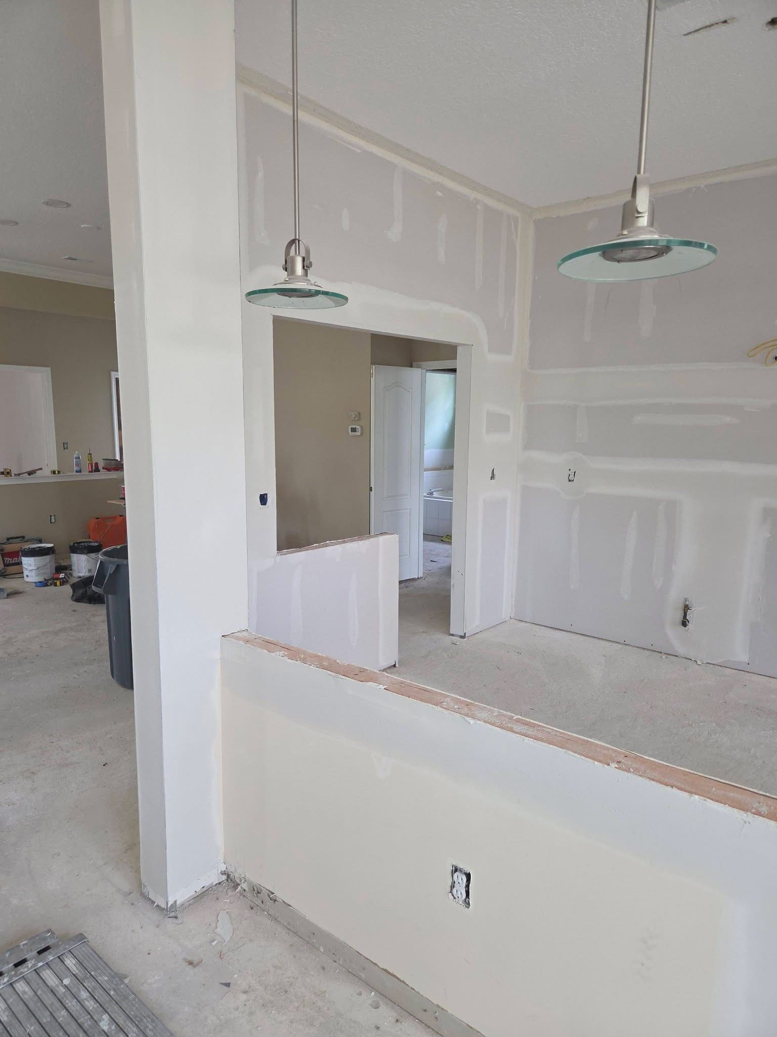 Partially finished kitchen with white walls, hanging light fixtures, and a pass-through counter opening