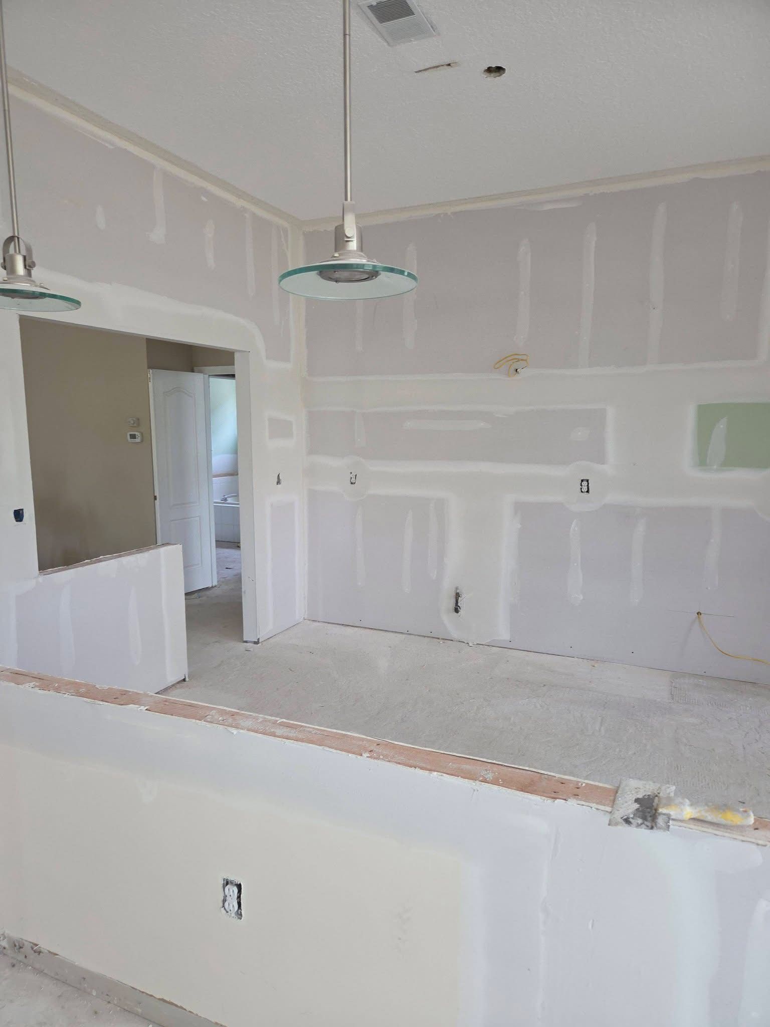 Partially renovated kitchen with drywall, hanging light fixture, and unfinished white cabinets